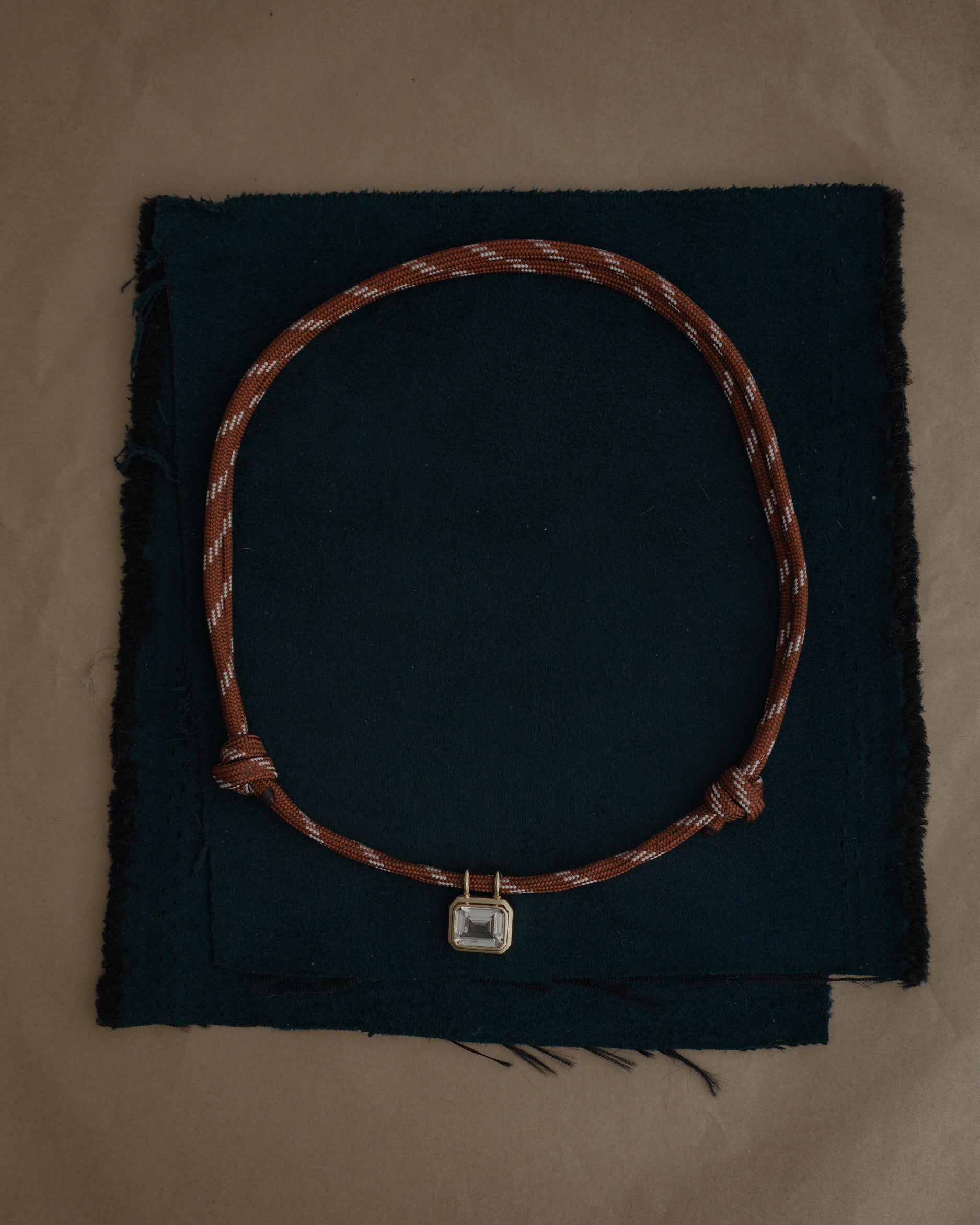 The Tommy Sapphire Pendant Necklace by Mod + Jo, featuring an adjustable brown paracord and a rectangular silver pendant, is displayed on two stacked pieces of dark fabric set atop a brown surface.