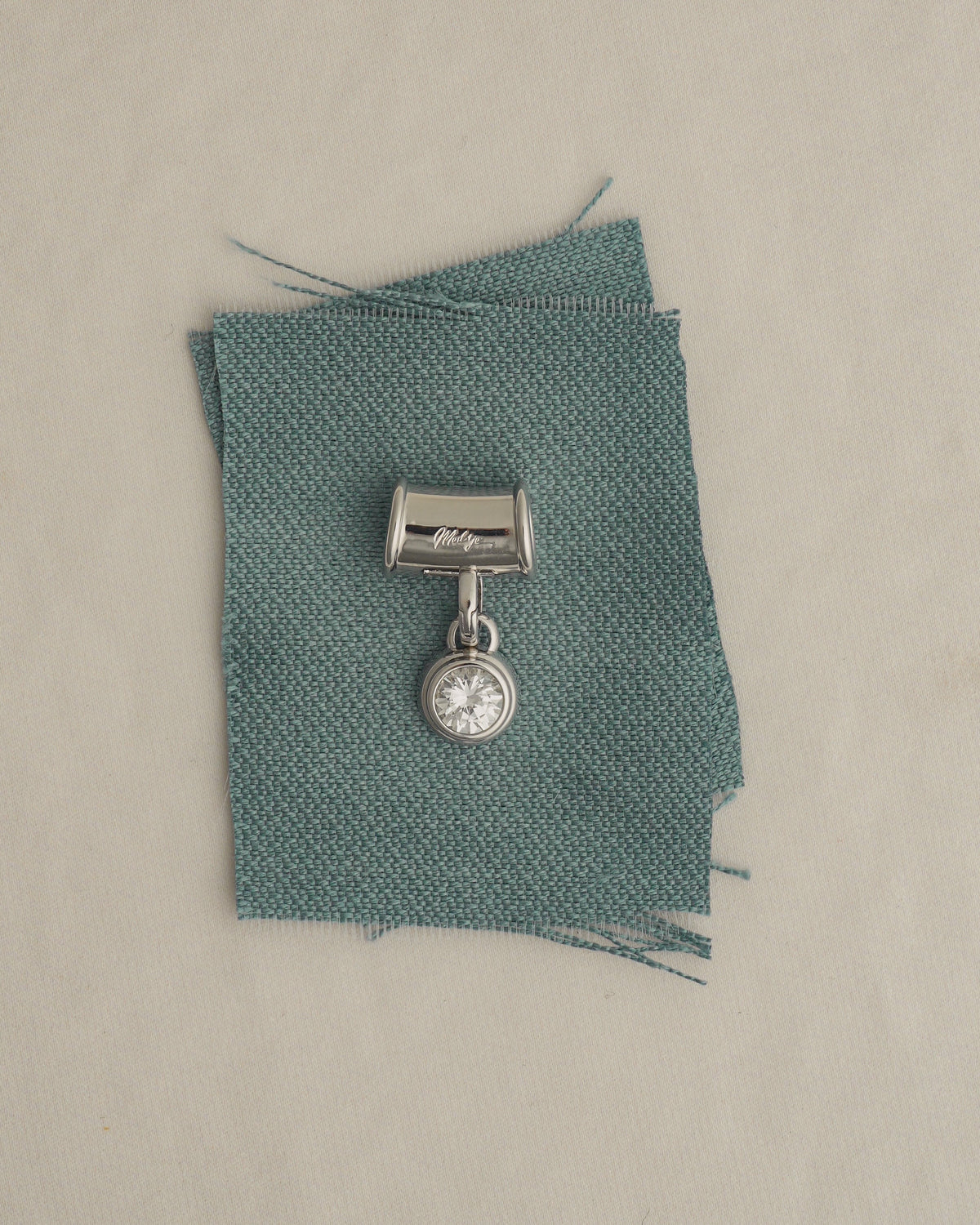 A nickel- and lead-free silver Tash Charm Enhancer featuring a single round stone, resting on a square of teal fabric.