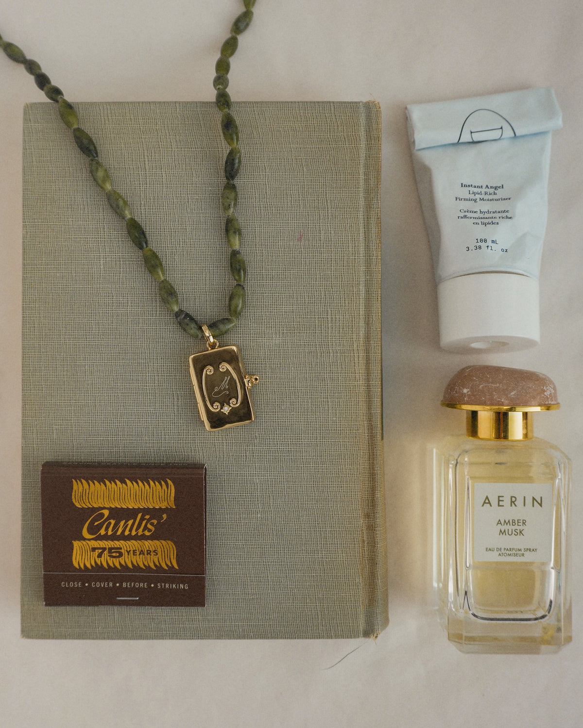 An 18″ Sutton Gemstone Necklace of green serpentine jade beads with a gold rectangular pendant, displayed on a book next to a tube of moisturizer, a perfume bottle, and a box of matches.