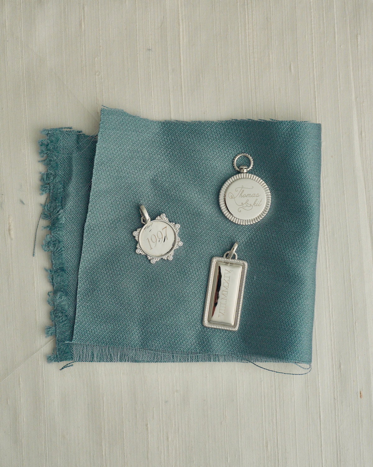 Three silver Hattie Engravable Charm tags engraved with 1997, Thomas, and Roman numerals, arranged as a tablet set on a piece of blue fabric.