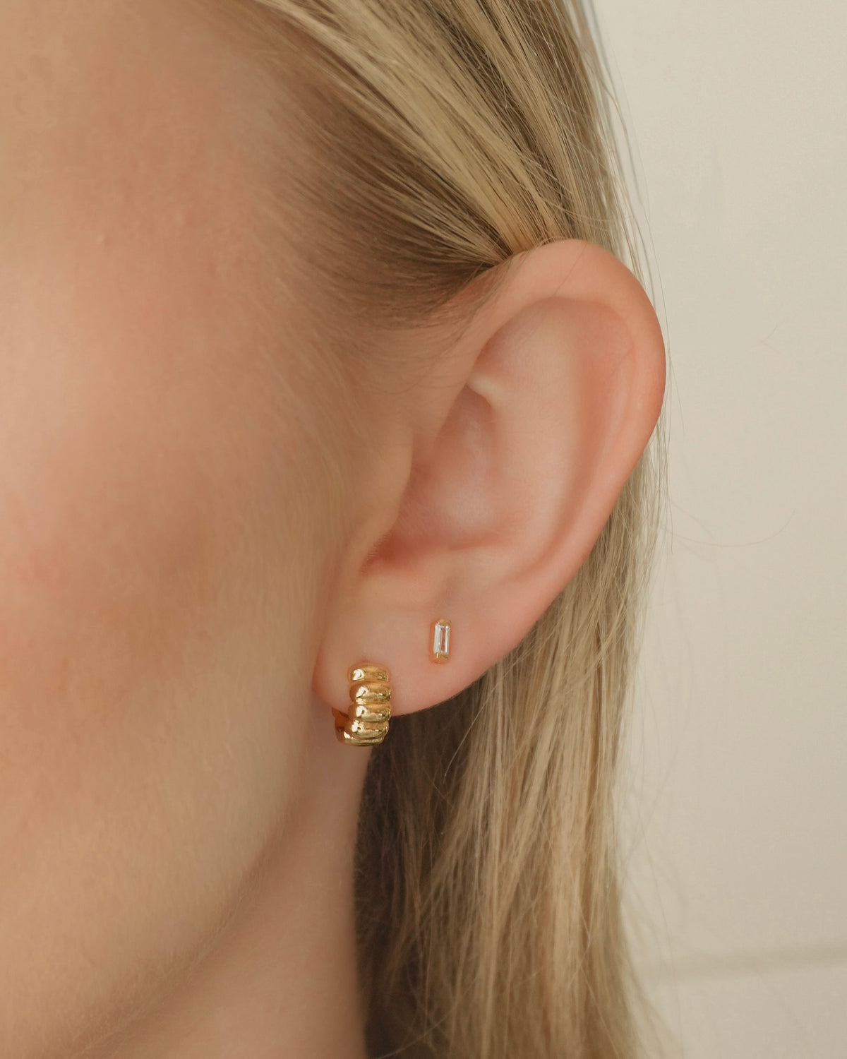 The Sylvie + Jadey Set features a gold Sylvie Huggie with a ribbed texture in the lower piercing and a small gold baguette stud with a rectangular stone in the upper piercing of an earlobe.