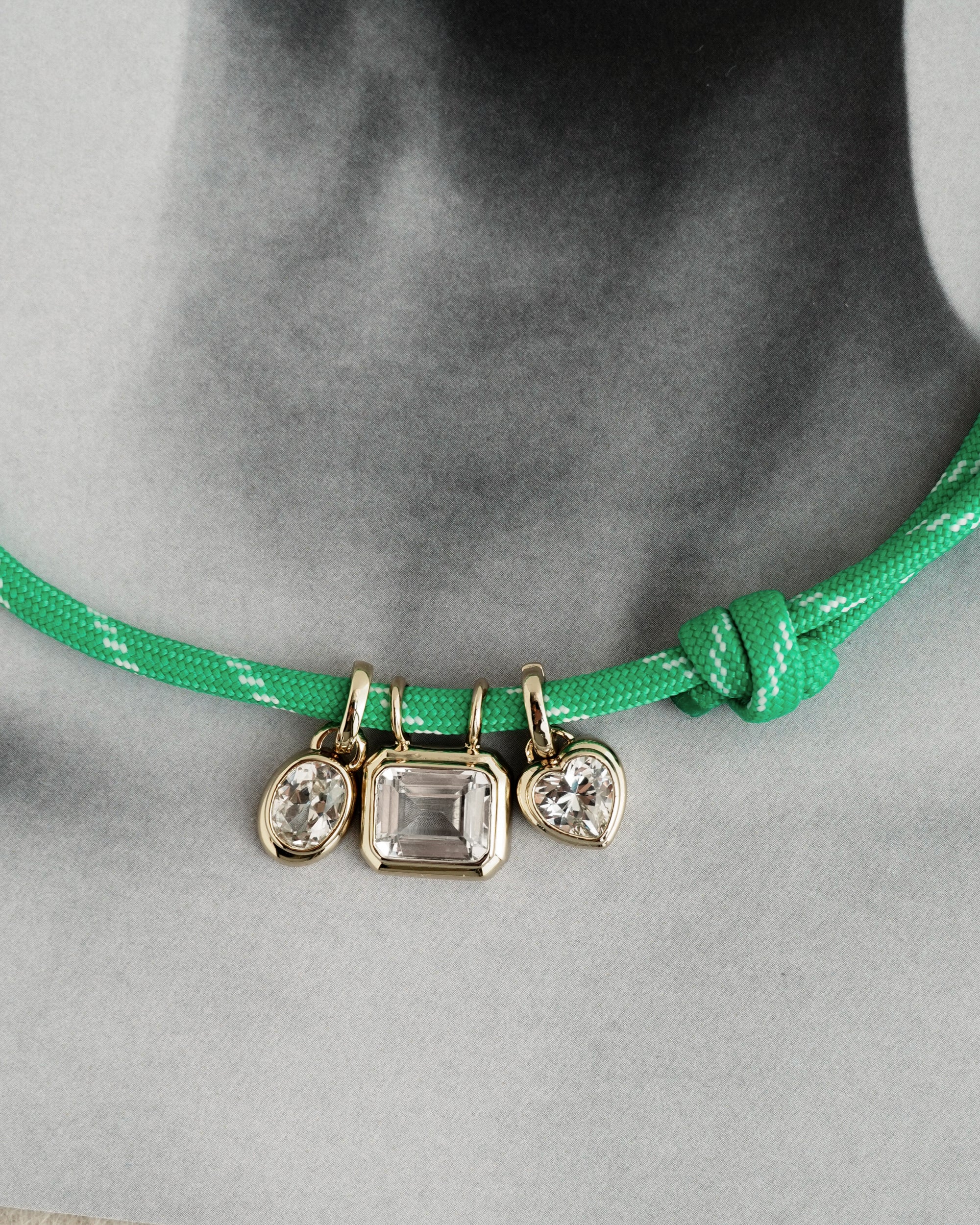 A green cord necklace with a knot, featuring three Mod + Jo Elsie Sapphire Bezel Charms crafted in 14k gold and set with clear lab-grown gemstones, displayed against a black and white photo background of a person’s neck and shoulders.
