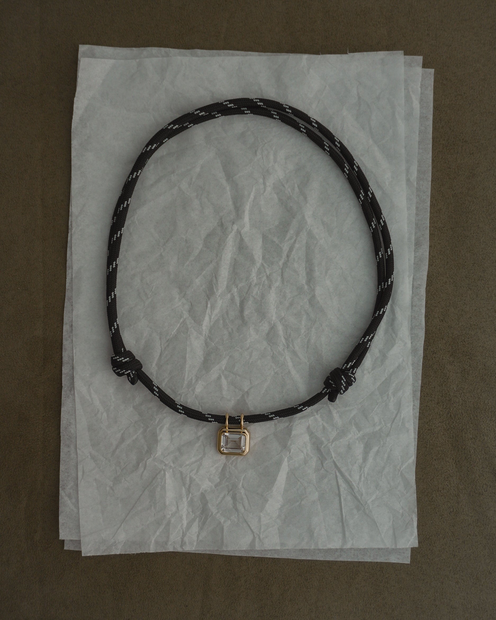 The Tommy Sapphire Pendant Necklace by Mod + Jo, featuring a black adjustable paracord with white specks and a small rectangular metal pendant, is displayed on crumpled tissue paper atop a brown surface.