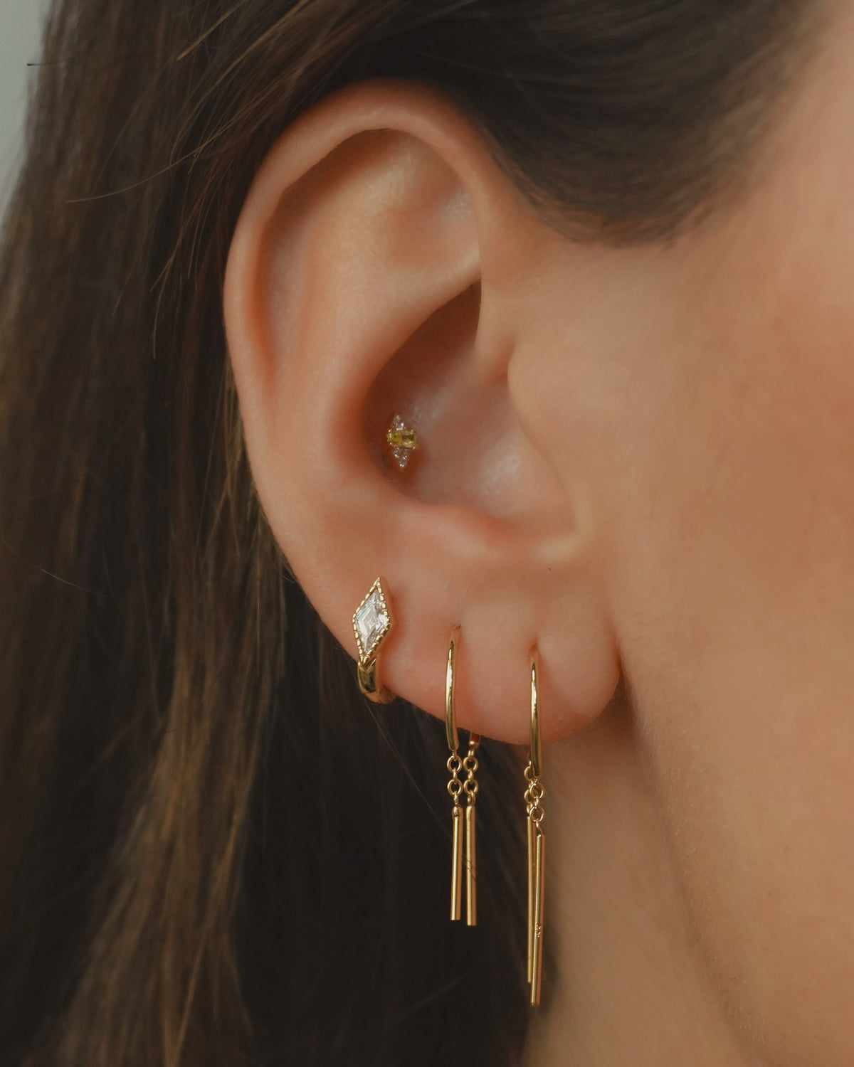 Close-up of a person’s ear adorned with four gold earrings: a small stud in the upper ear, a marquise-shaped stud in the lobe, and two Mod + Jo Mila Smooth Threader Huggies—14k gold hoops with dangling bars—in the lower lobe. This elegant arrangement features hypoallergenic jewelry, making it perfect for sensitive skin.