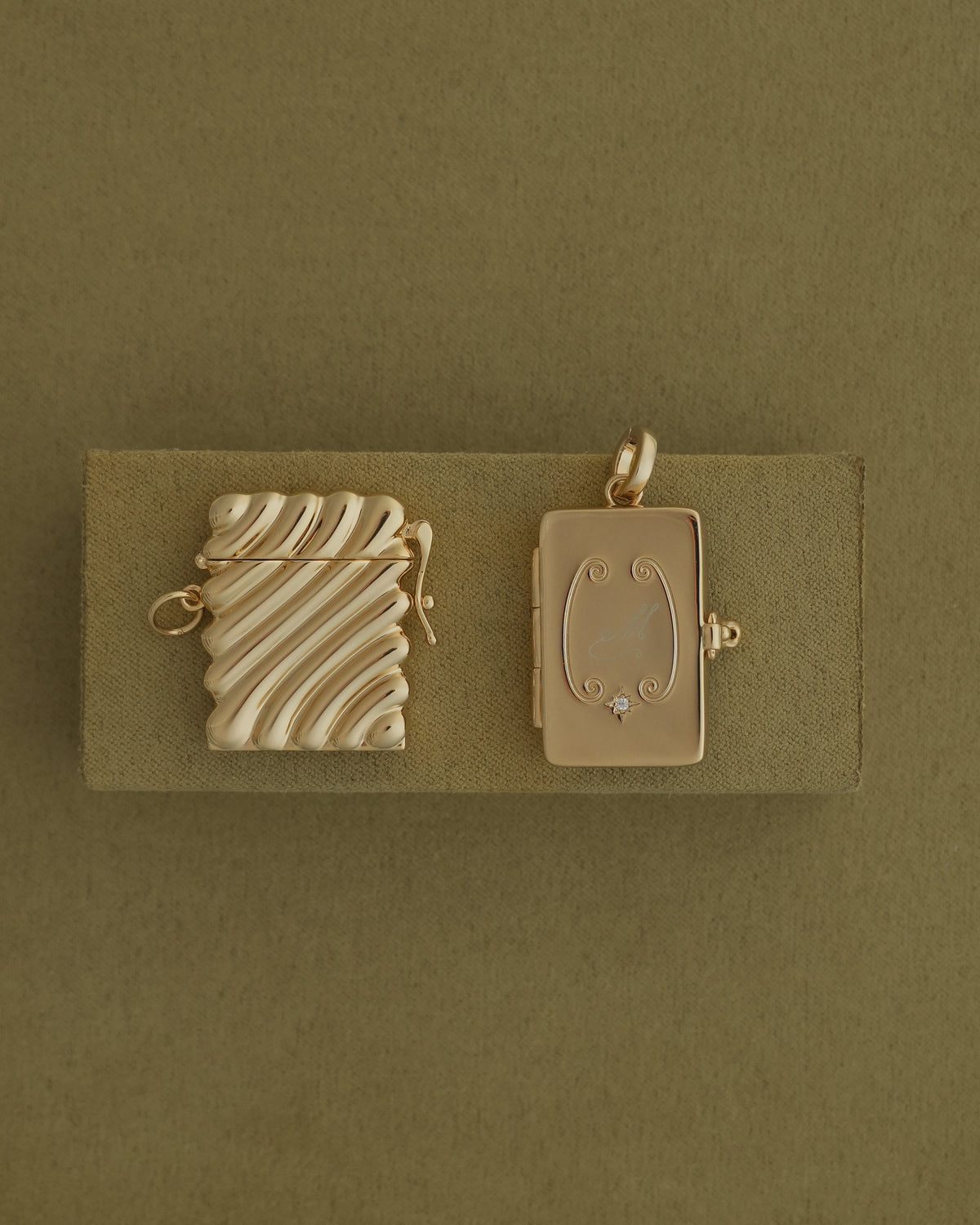 Two gold-plated Caspia Vesta Case Charm rectangular charms rest on a textured olive green base, one with a wavy ridged surface and the other featuring an engraved scrollwork design with a small inset gemstone.