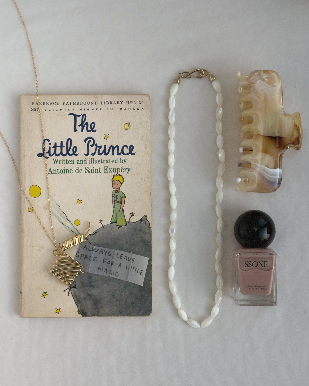 A copy of The Little Prince book next to a Caspia Vesta Case Charm pendant, a white beaded necklace, a hair claw clip, and a bottle of pink nail polish.
