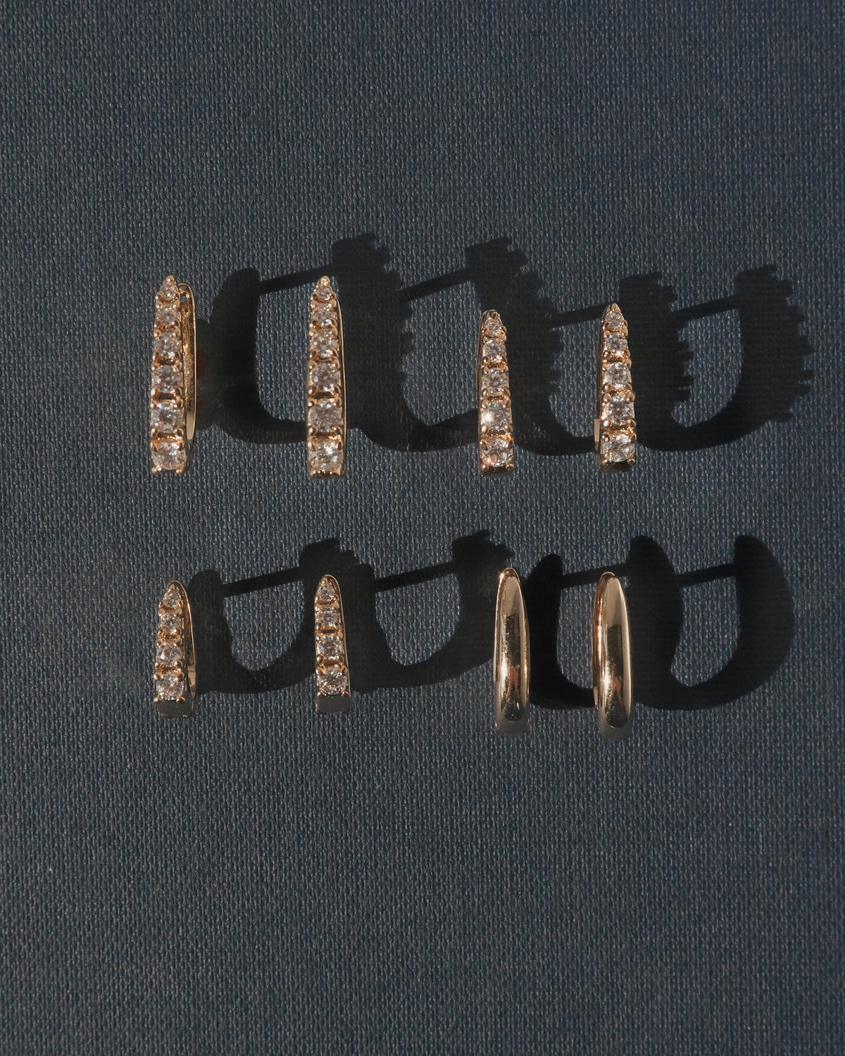 Six Jules Smooth Huggies gold hoop earrings, some featuring a row of embedded stones, arranged in two rows on a dark textured background and crafted with advanced plating technology.