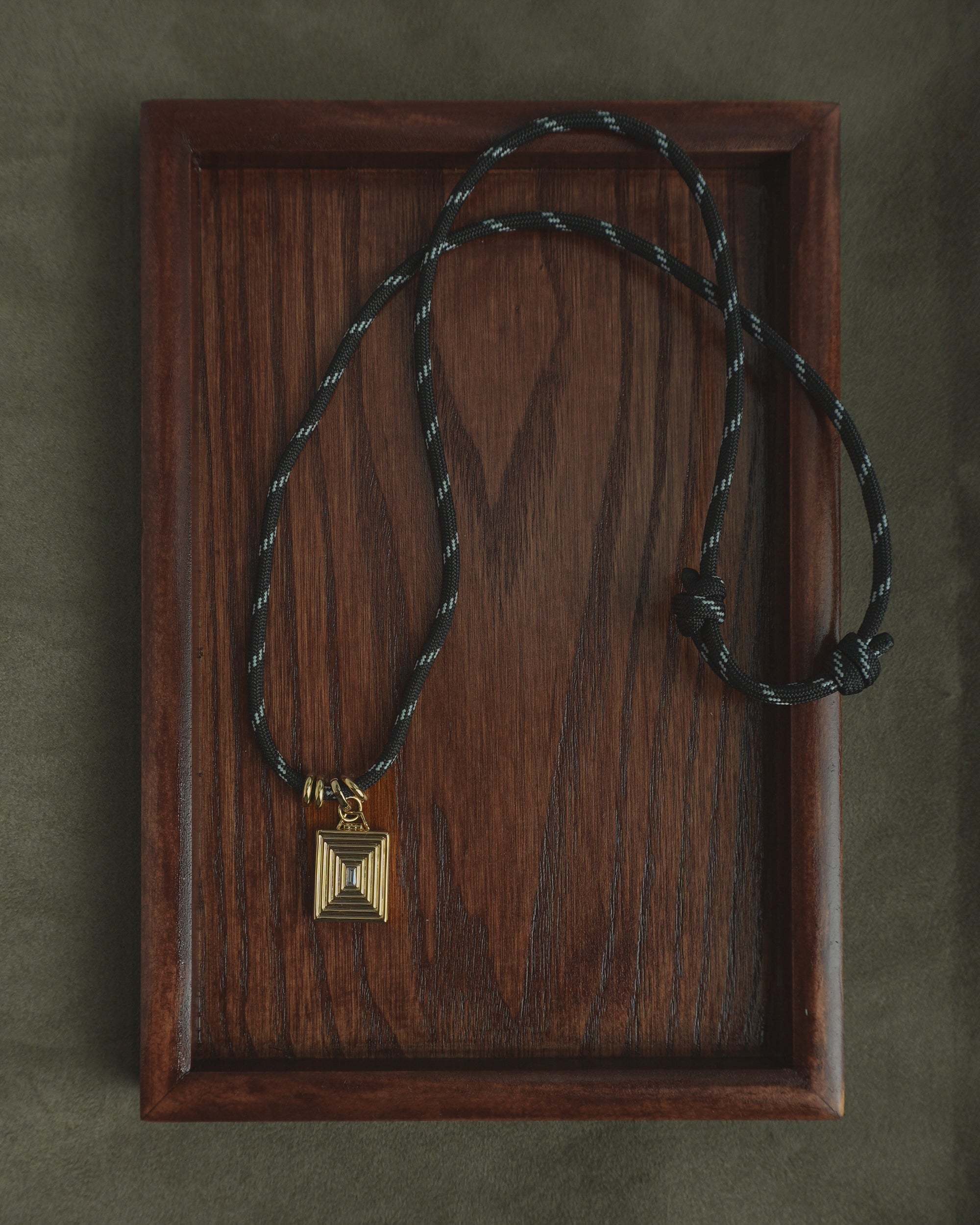 The Tommy Cord Necklace by Mod + Jo features a gold geometric pendant suspended from an adjustable black paracord with gold-plated spacers, all elegantly displayed on a rectangular dark wood grain tray.