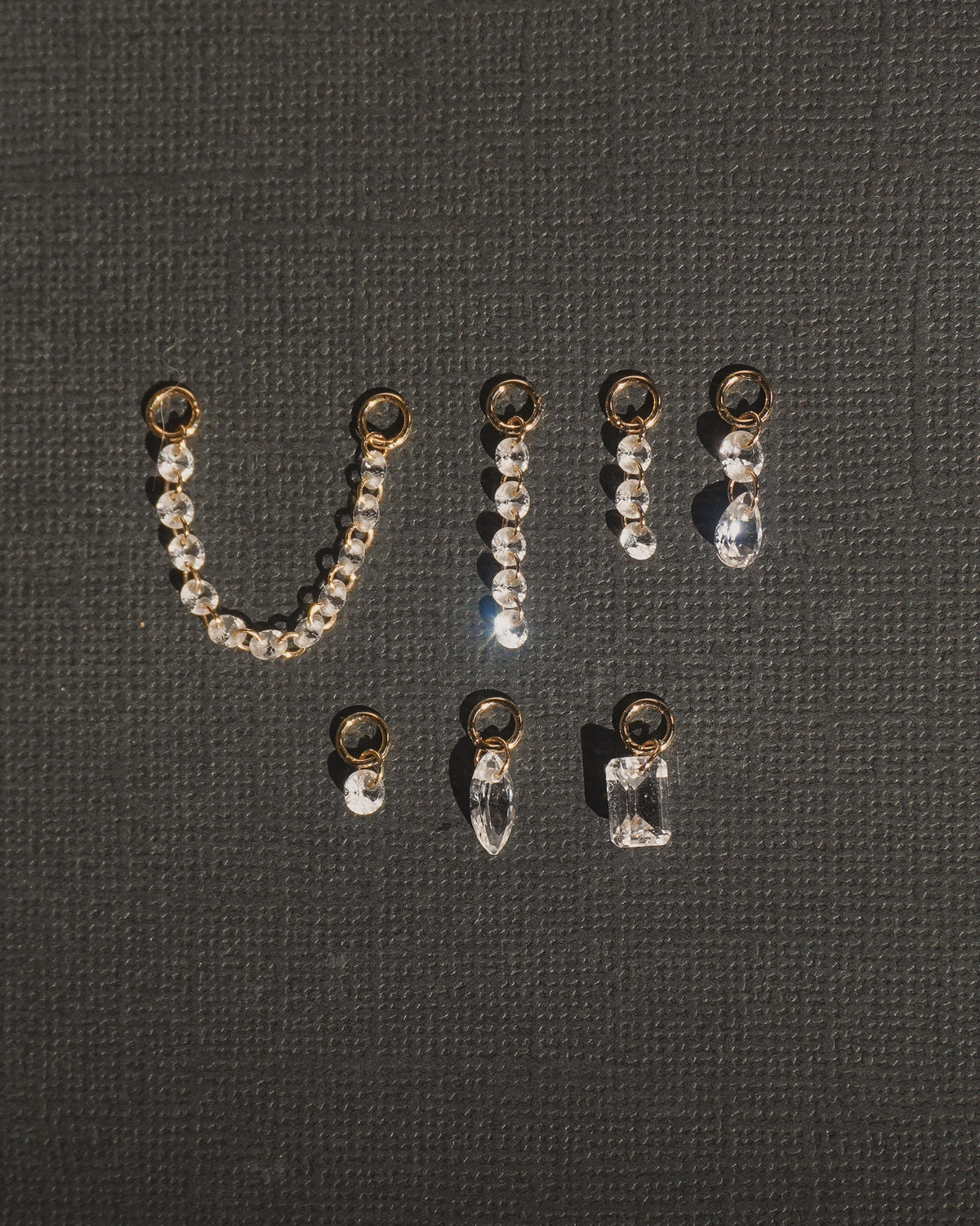 Seven Mod + Jo Noa Sapphire Earring Charms in gold-plated metal featuring various lab-grown white sapphire shapes including chains, drops, and geometric solitaires.