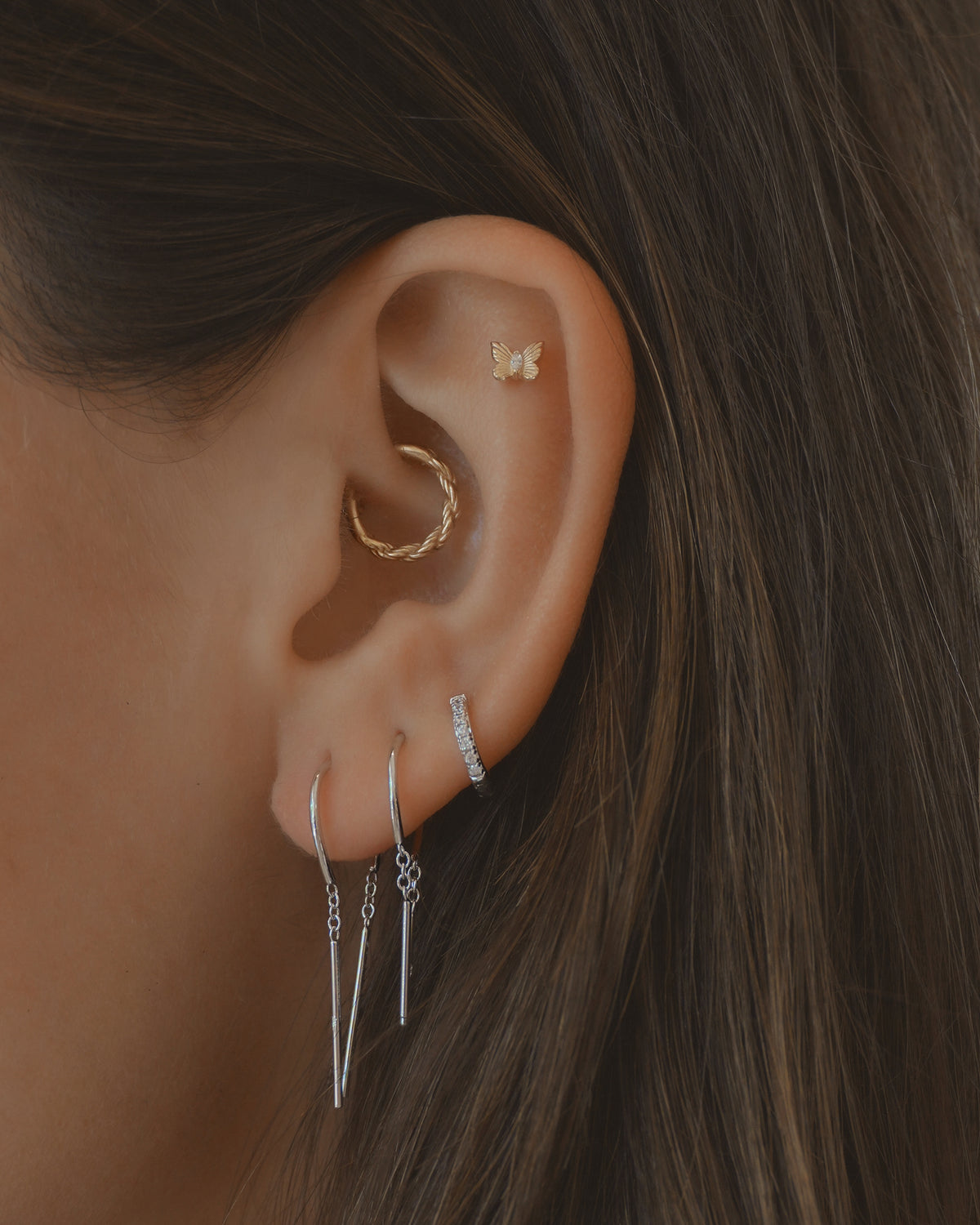 A close-up of an ear adorned with four earrings: a gold twisted hoop on the upper ear, a small butterfly stud above it, and two silver drop earrings plus one silver hoop earring with stones on the lower lobe. Featured among them are the Mila Smooth Threader Huggies by Mod + Jo. All pieces are nickel- and lead-free for comfortable wear. Medium brown hair frames the ear.