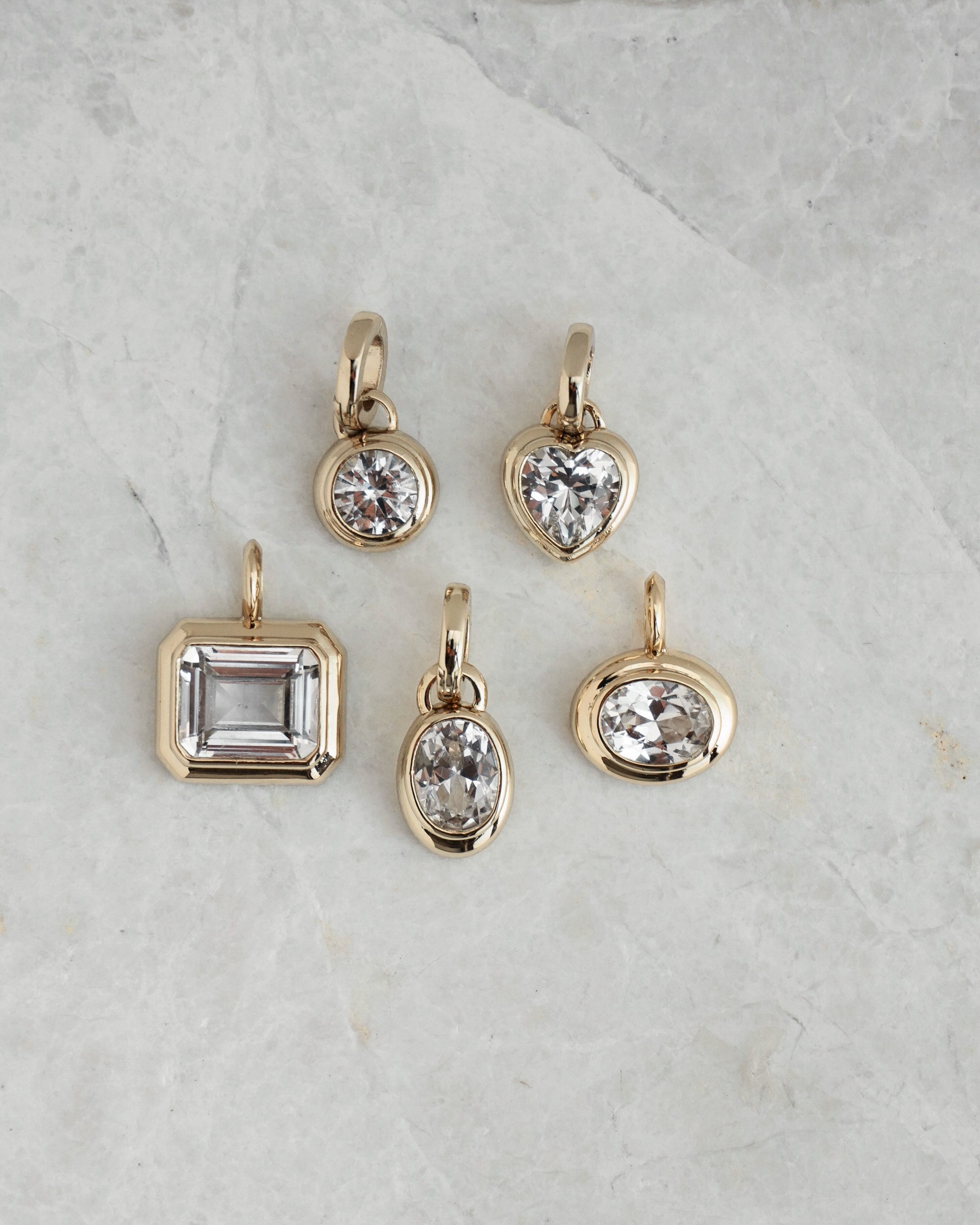 Five Mod + Jo *Preorder* Tove Sapphire Bezel Charms, featuring lab-grown sapphires in round, heart, rectangular, and oval shapes set in 14k gold, are displayed on a light gray marble surface.