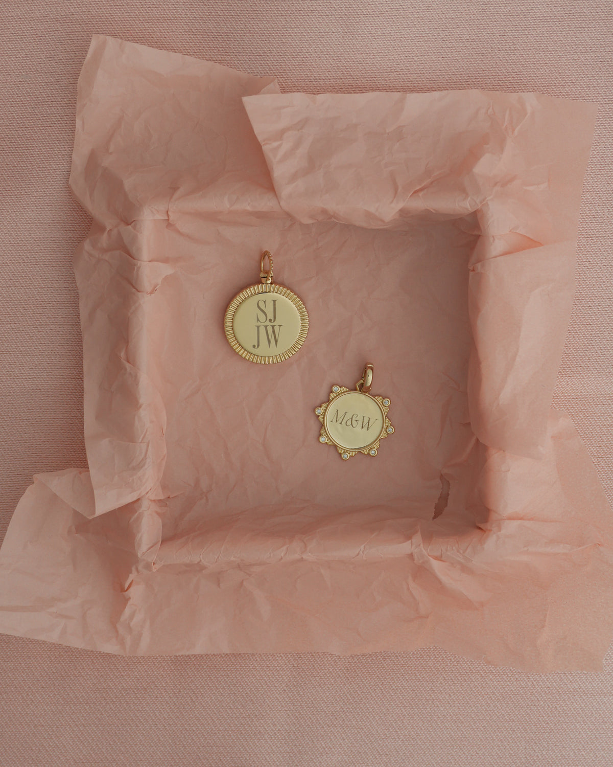 Two gold-plated Gia Engravable Charm pieces in a pink tissue-lined box: one coin-edge disc engraved with SJ JW, and one scalloped-edge charm inset with crystals engraved with M&amp;W.