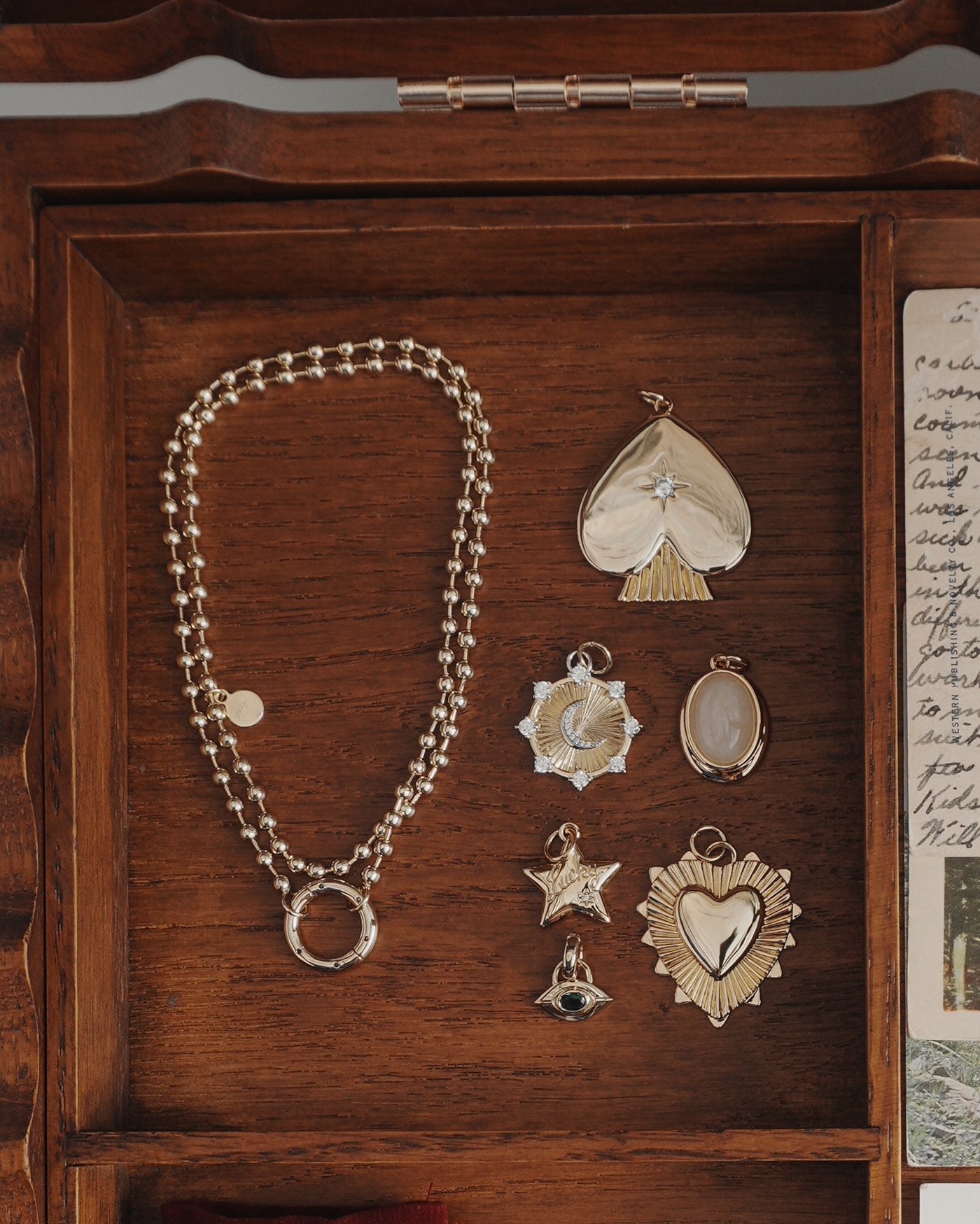 The Rory Gemstone Charm set by Mod + Jo features a lustrous gold chain necklace adorned with six unique 14k gold pendants—including heart shapes, stars, and an eye—beautifully presented in a wooden jewelry box beside a handwritten note. This stunning collection combines timeless elegance with meaningful symbols of self-love, making it the perfect gift or an everyday treasure to cherish for yourself.