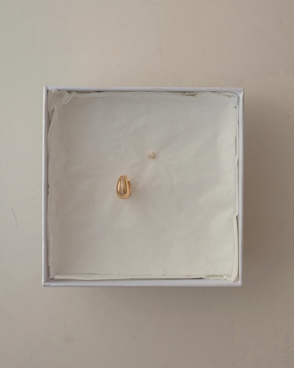 A Brianne + Ace Set featuring a small gold chunky huggie encrusted with crystals and a matching single stud earring resting inside a white gift box lined with tissue paper.