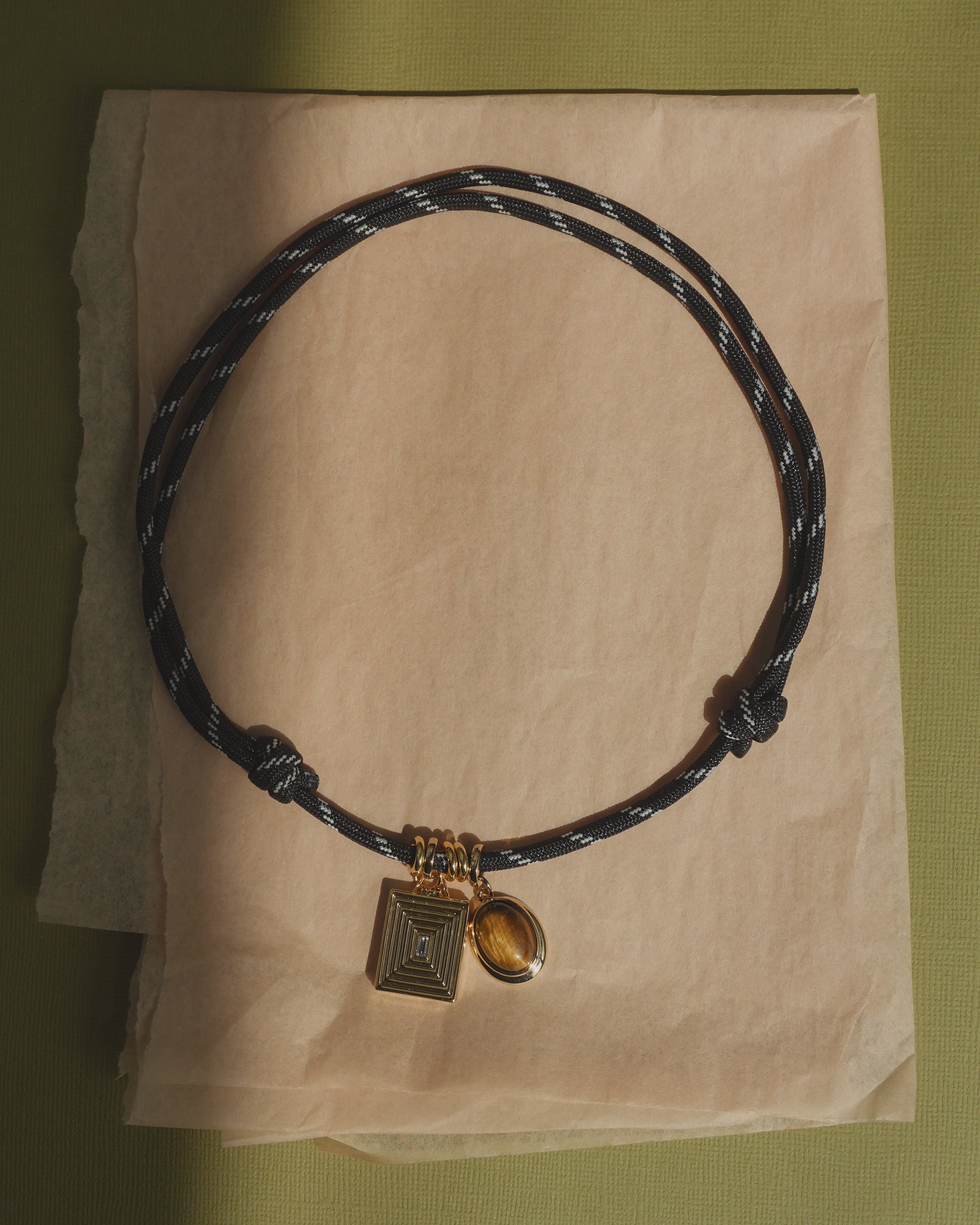 The Tommy Cord Necklace by Mod + Jo features a black and white braided paracord adorned with a gold rectangular charm, a round tigers eye charm, and gold-plated spacers, artfully displayed on crumpled tan paper over a green surface.