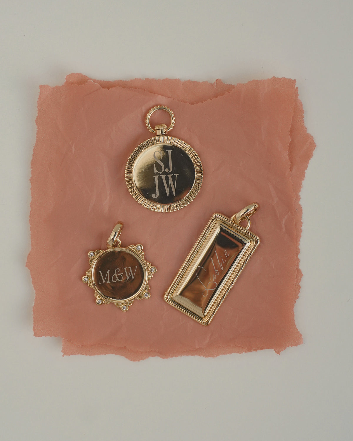 Three Remi Engravable Charm pieces on a pink paper background: a round charm marked S.J. JW, a circular cubic zirconia charm engraved with M&amp;W, and a rectangular charm engraved with Libbie.