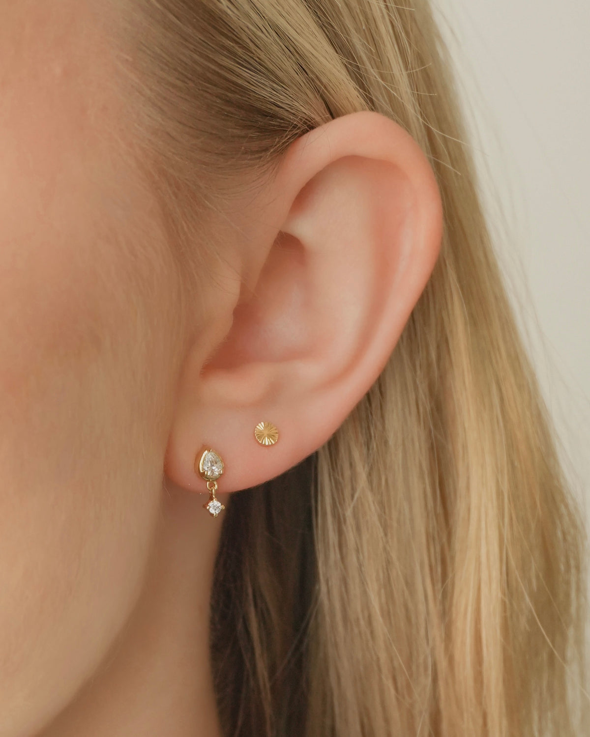 A person wearing the Quinn + Dela Set, featuring a small gold Quinn Stud in the upper piercing and a pear-shaped, gold Dela Fluted Stud with a dangling crystal in the lower piercing.