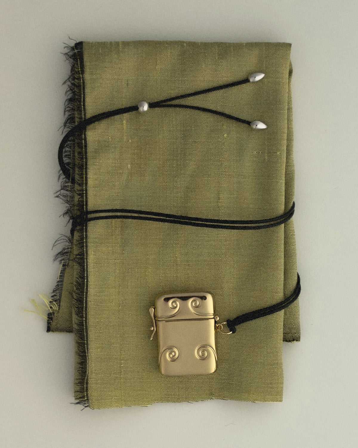 A gold-plated Victorian Sybil Vesta Case Charm with decorative swirls rests on a folded green fabric cloth, held by a black cord with silver accents.