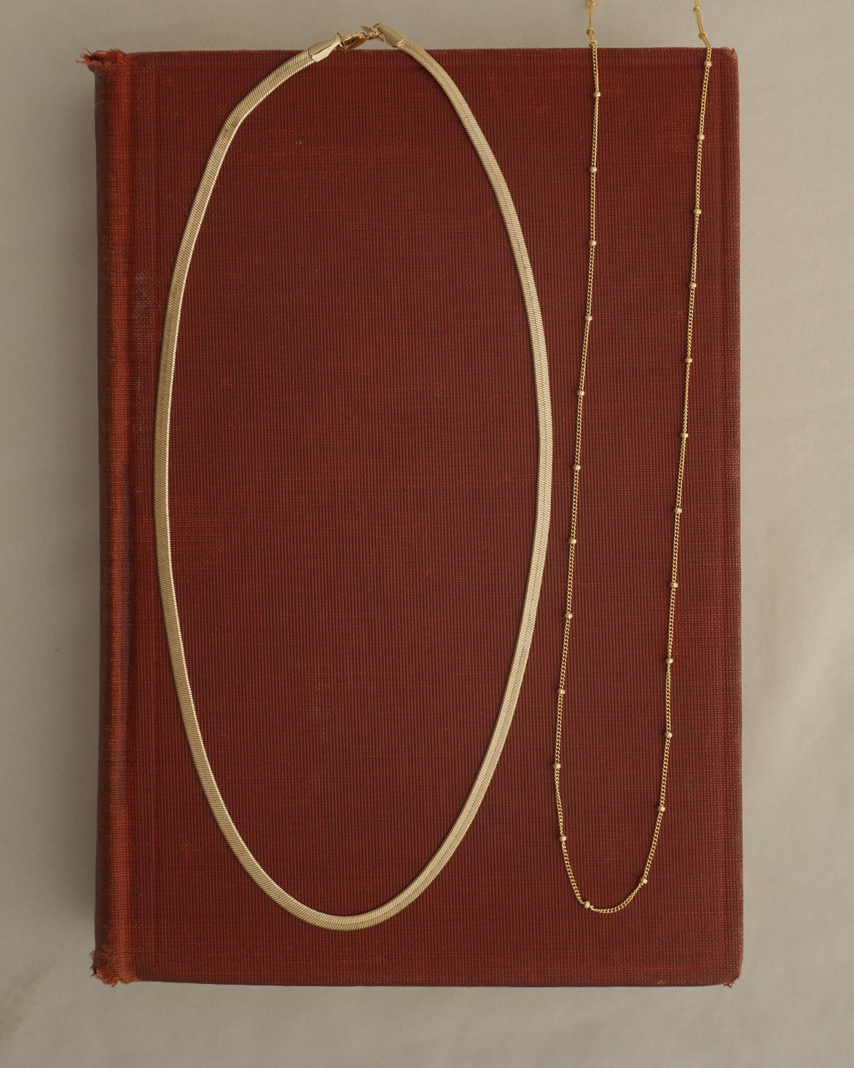 Mod + Jo Everyday Necklace Set featuring a gold herringbone necklace and a delicate gold satellite chain necklace with small beads, resting on a red book.