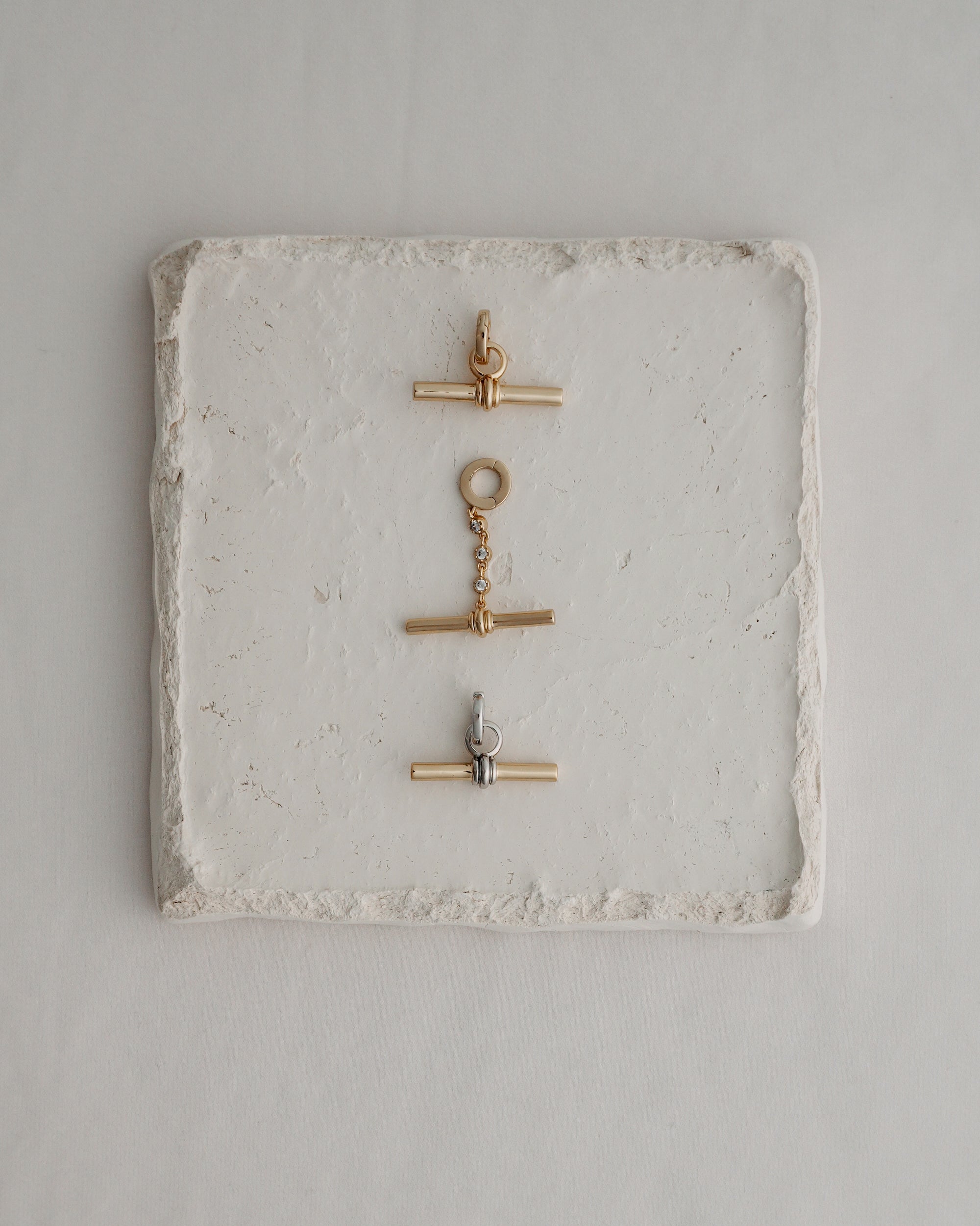 Three metal toggle clasps—two in gold and one in silver—are arranged vertically on a square, textured white stone tile set against a light background, echoing the elegant detail of Mod + Jo's Elliot Toggle Charm and the refined style characteristic of their 14k gold-inspired jewelry.