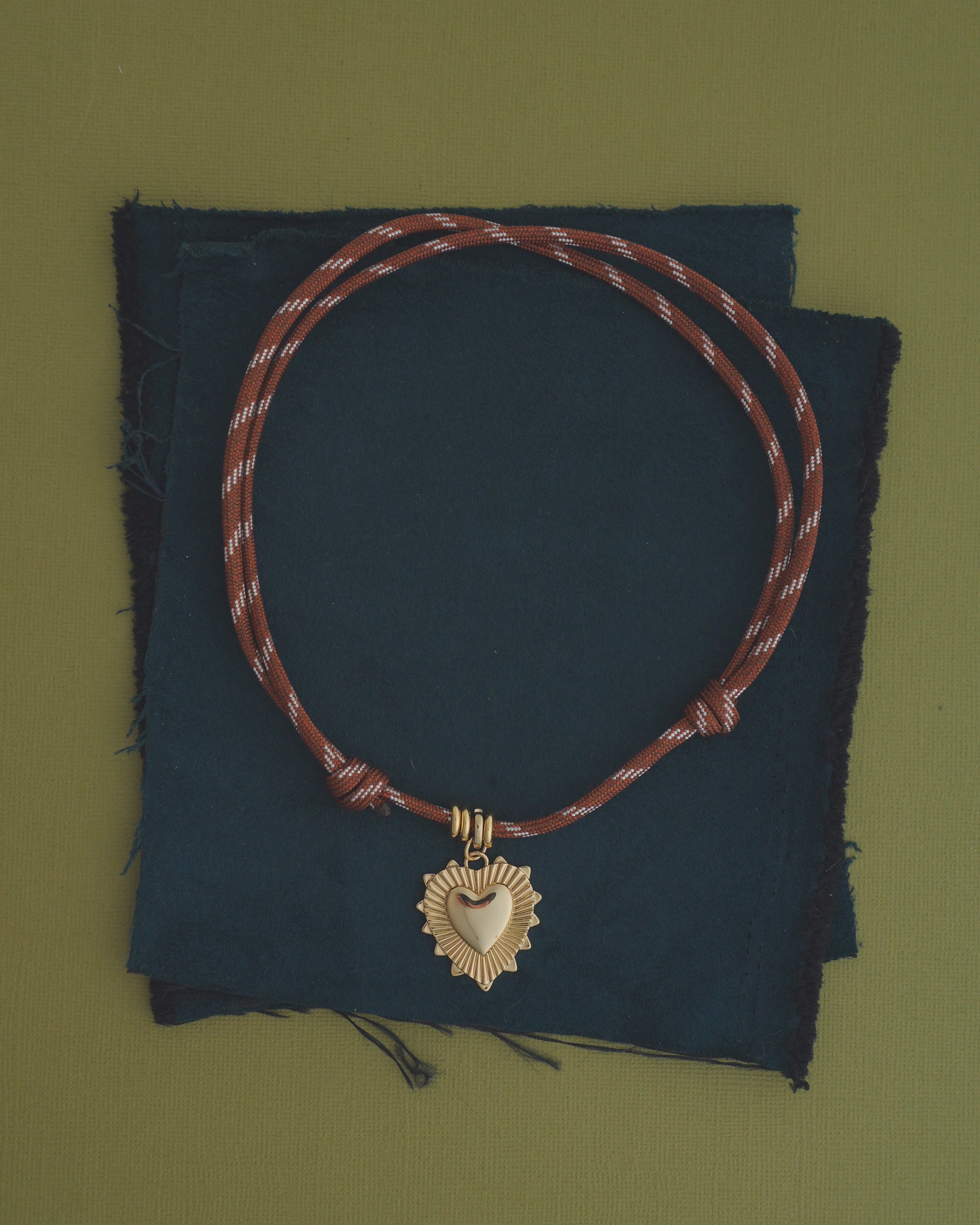 The Tommy Cord Necklace by Mod + Jo, featuring a brown paracord with a gold heart-shaped pendant and gold-plated spacers, is displayed resting on stacked dark blue fabric squares against a green background.