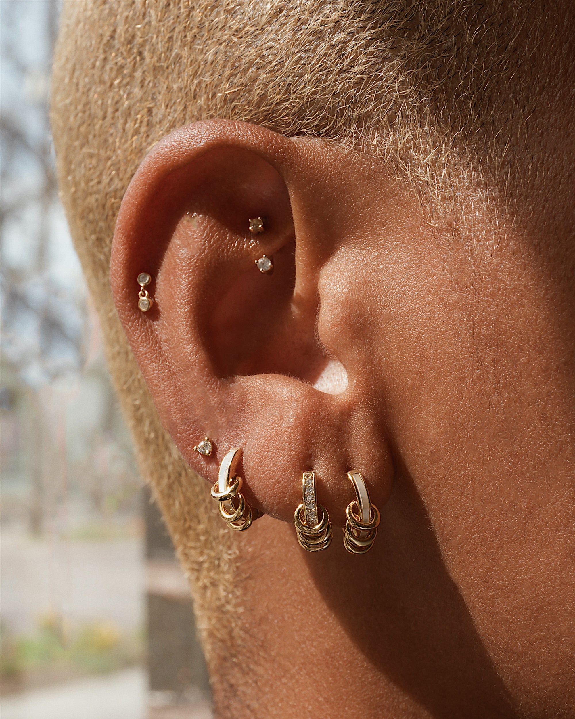 A close-up of a person’s ear with short hair, styled with multiple gold and diamond earrings, including Mod + Jo’s tarnish-resistant Levi Huggies on the lobe and cartilage.