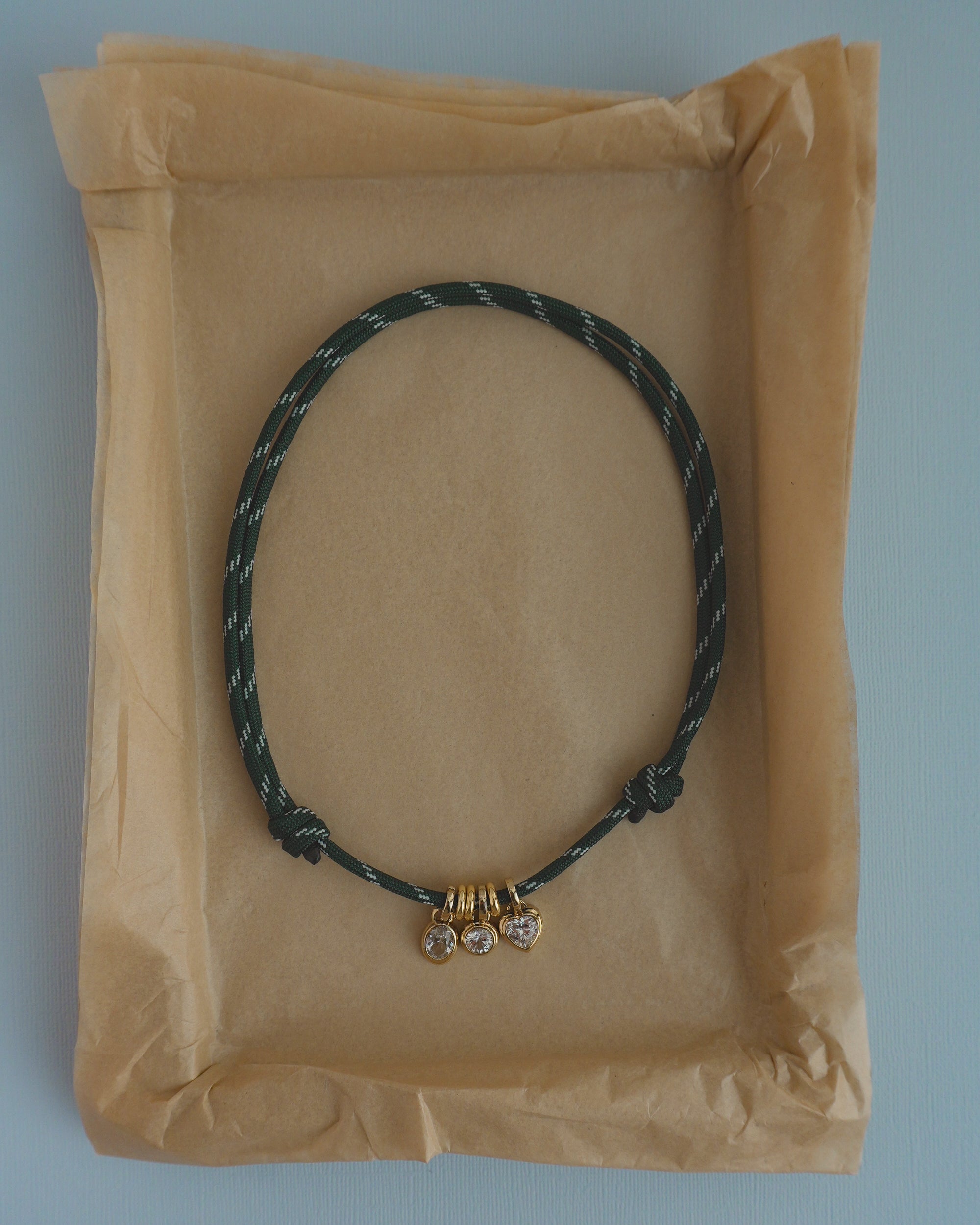 The Mod + Jo Tommy Cord Necklace features a green cord adorned with three small gold and crystal charms and gold-plated spacers, elegantly displayed on crumpled brown paper in a shallow rectangular box.