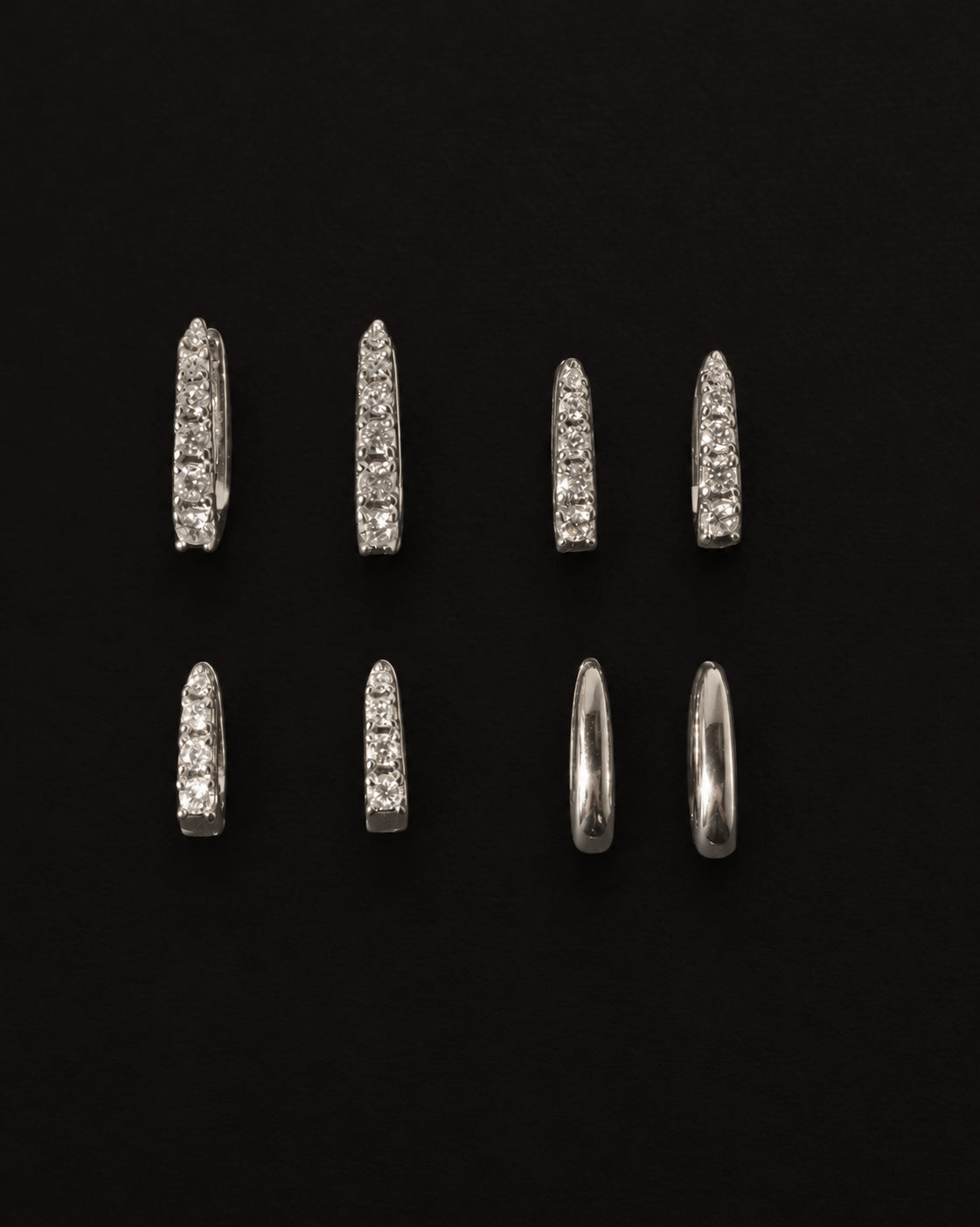 Eight nickel- and lead-free Jules Smooth Huggies arranged in two rows against a black background, with some featuring embedded stones and others with a plain polished finish.
