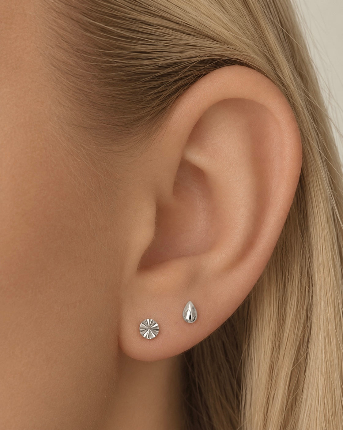 Close-up of an ear wearing the Dela + Uma Set, featuring a small, fluted stud earring and a smooth, silver teardrop stud earring.