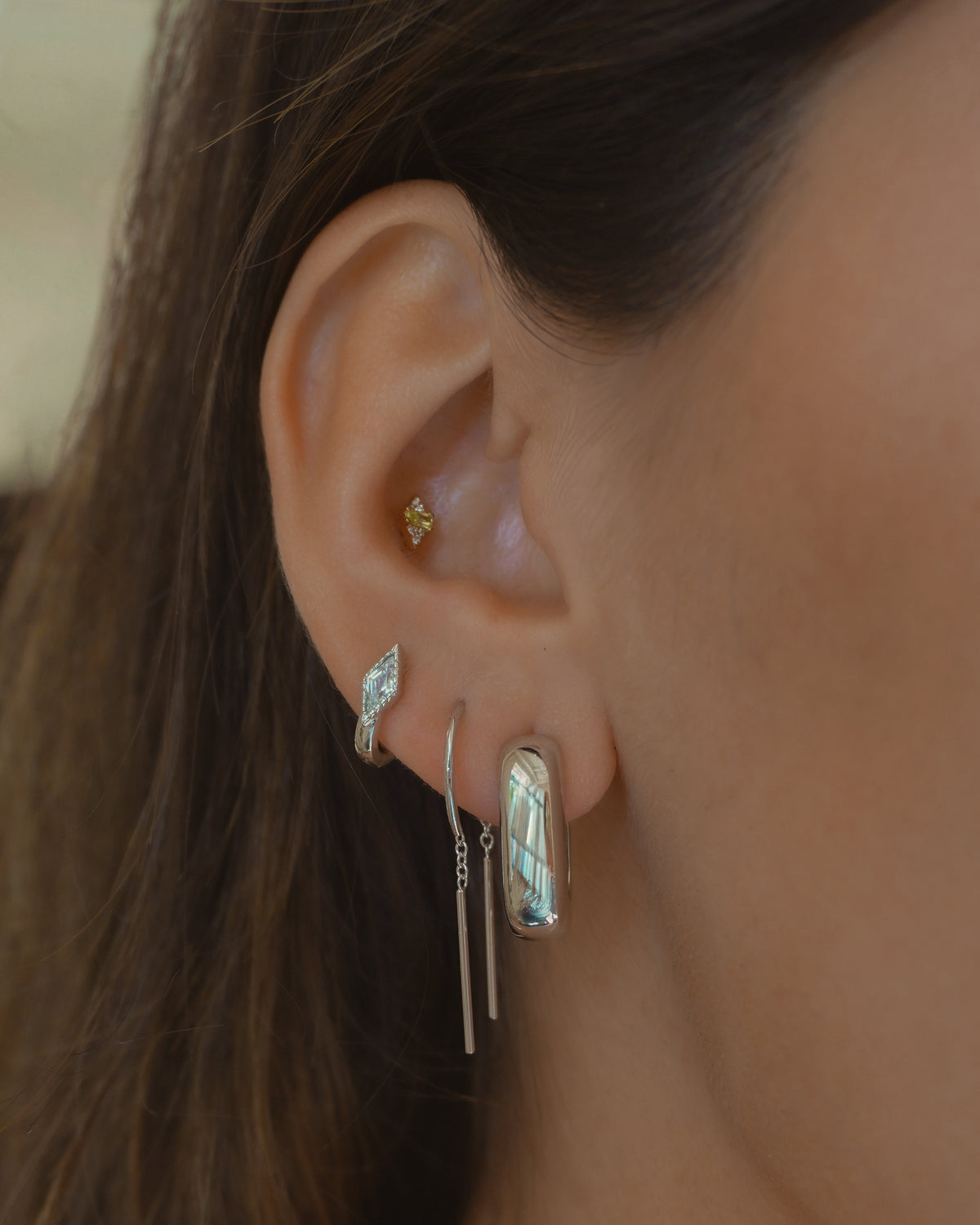 A close-up of a person’s ear showcases four earrings, including the Mila Smooth Threader Huggies from Mod + Jo: a small yellow stud, a curved silver piece, a dangling bar earring, and a large rectangular design with an iridescent inlay—each crafted in rhodium plate for extra shine. This curated look complements classic hoop styles or sparkling cubic zirconia accents.