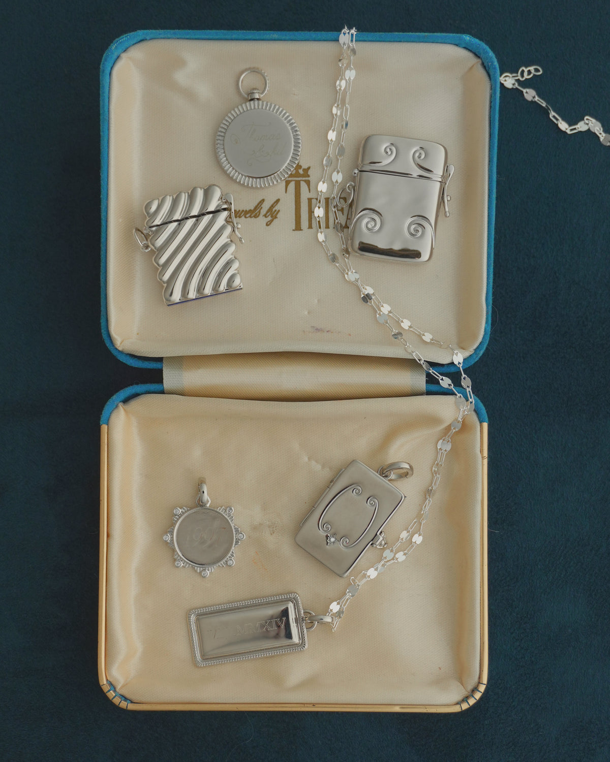 Five silver-plate charms, including two Caspia Vesta Case Charm pendants, a circular pendant, an ornate rectangular pendant, and a rectangular bar pendant with Roman numerals, presented in an open velvet-lined jewelry box with a silver chain.