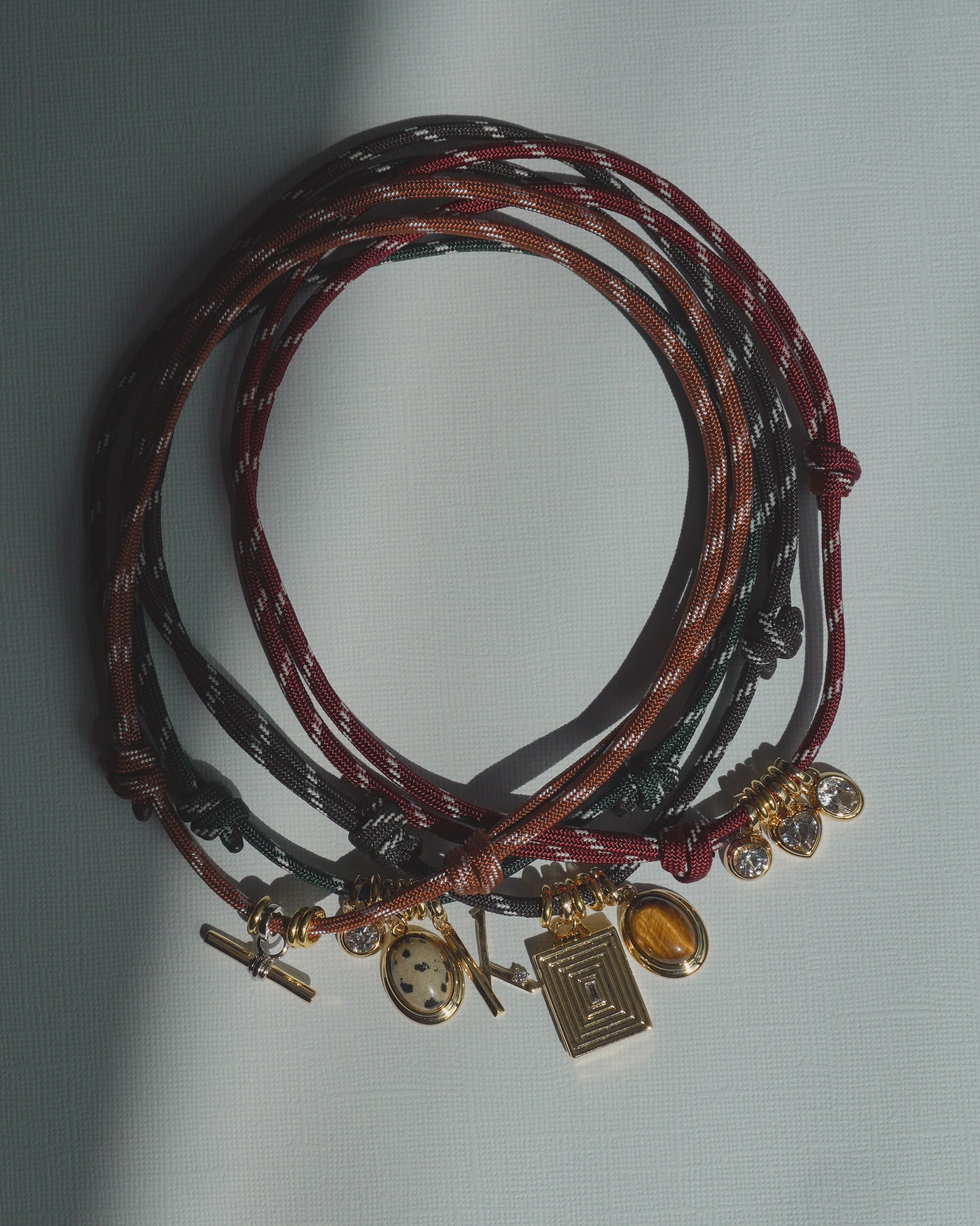 Three multicolored Tommy Cord Necklaces by Mod + Jo, adorned with gold and gemstone charms, are arranged in overlapping circles on a light textured surface with sunlight casting delicate shadows. Gold-plated spacers add an elegant touch to each necklace, enhancing the vibrant look of these lively accessories.