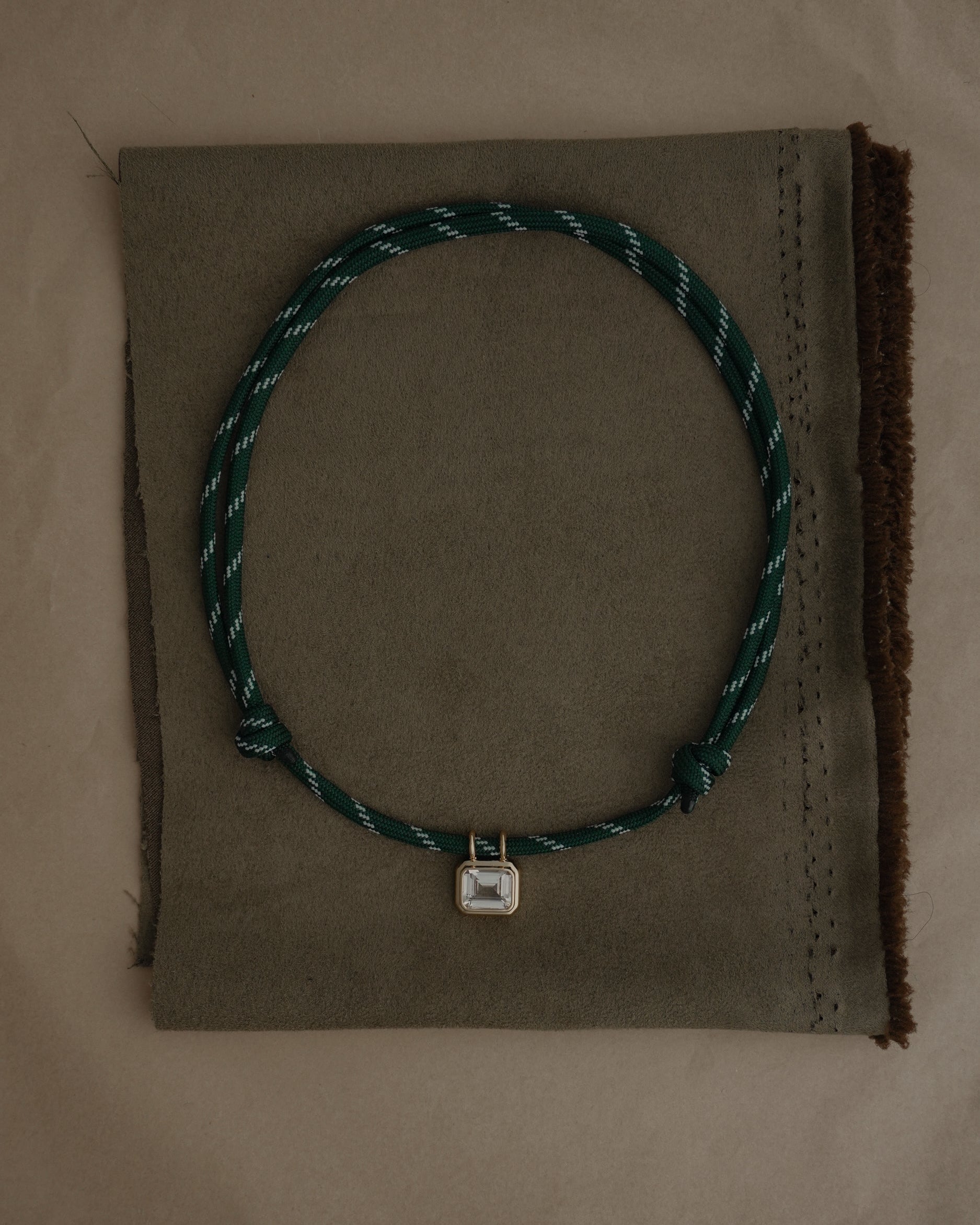 The Mod + Jo Tommy Sapphire Pendant Necklace features an adjustable green and white paracord with a small square gold bezel-set sapphire, showcased on a folded piece of brown fabric.