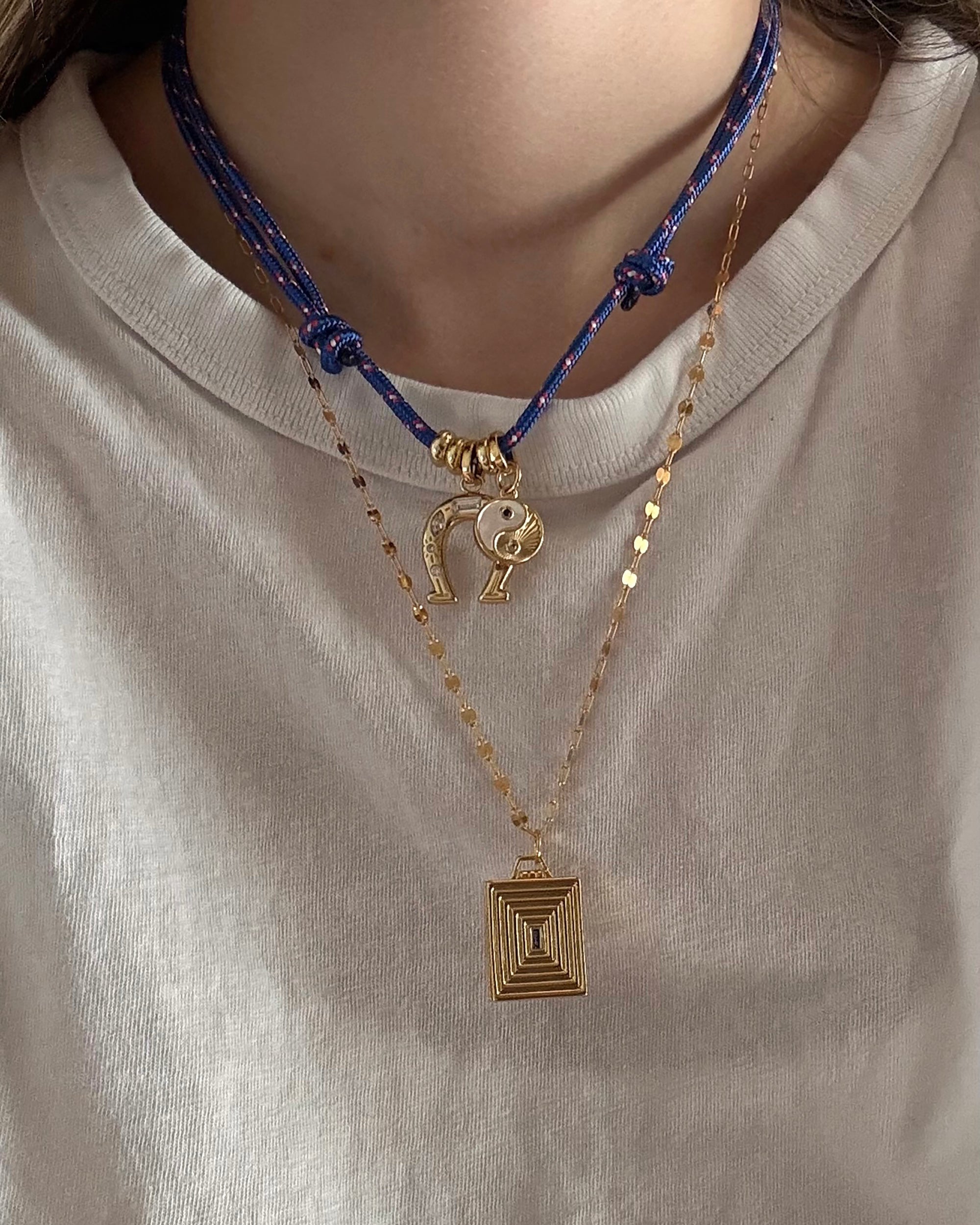 A person wears a white shirt and two necklaces: the Mod + Jo Tommy Cord Necklace, an adjustable blue paracord with gold charms, including a smiley face, and a gold plated necklace with a rectangular geometric pendant.