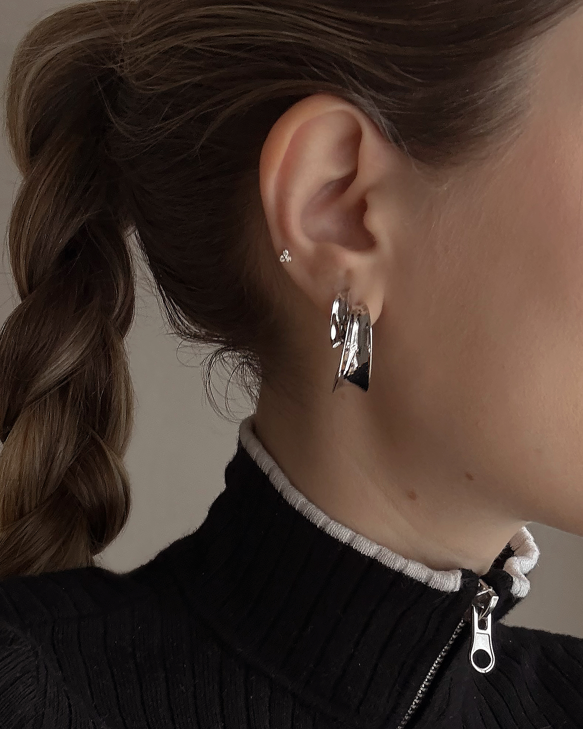 Close-up of a person with braided brown hair wearing a black ribbed zip-up top and Mod + Jo's Grande Valen Tapered Hoops—large, shiny, rhodium-plated earrings—in one ear. The image shows part of their face and neck.
