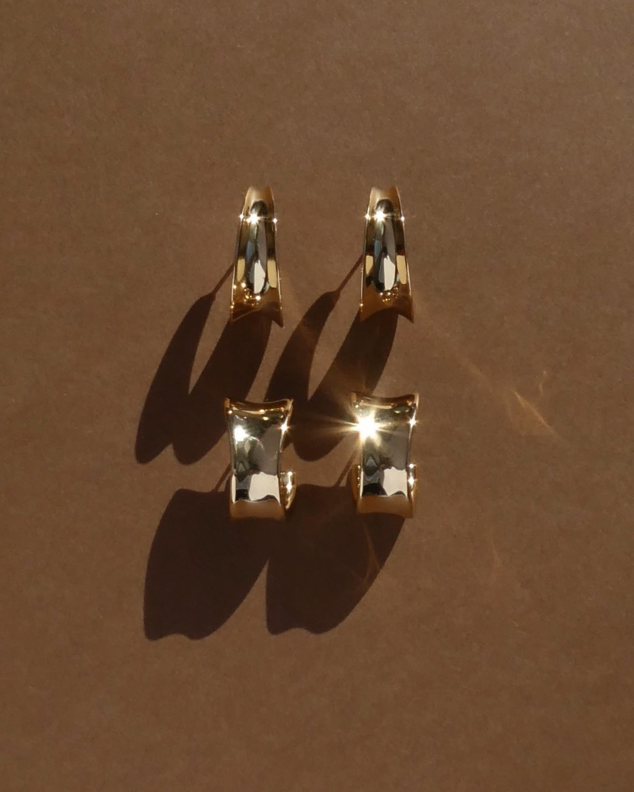 Four shiny Grande Valen Tapered Hoops by Mod + Jo, 14k gold plated and nickel free, are arranged in two rows on a brown surface, casting soft shadows as light creates a bright glare on the lower gold hoops.