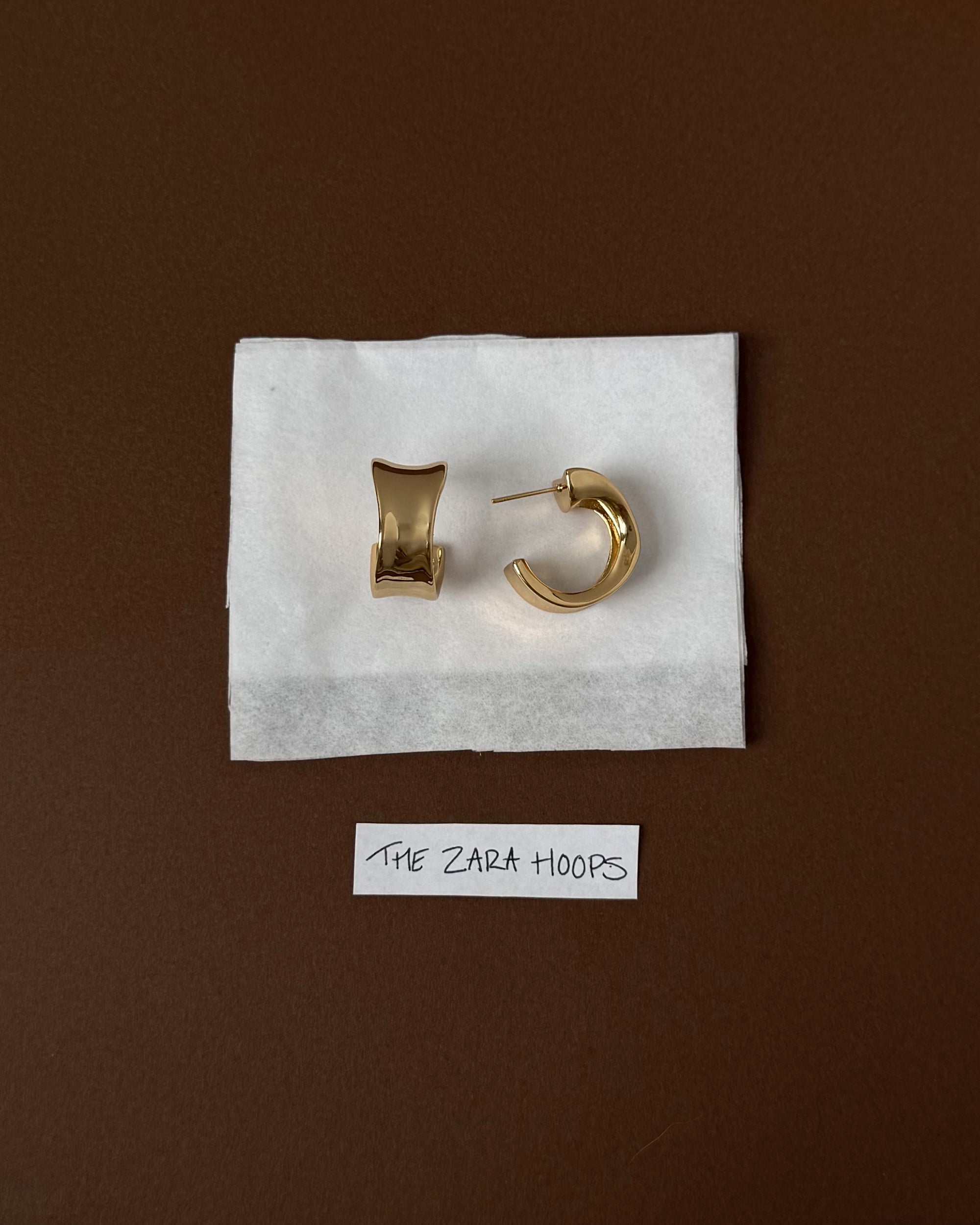 A rectangular gold earring and a circular gold hoop earring are displayed on a white cloth with a handwritten label below reading THE ZARA HOOPS, all set against a brown background.