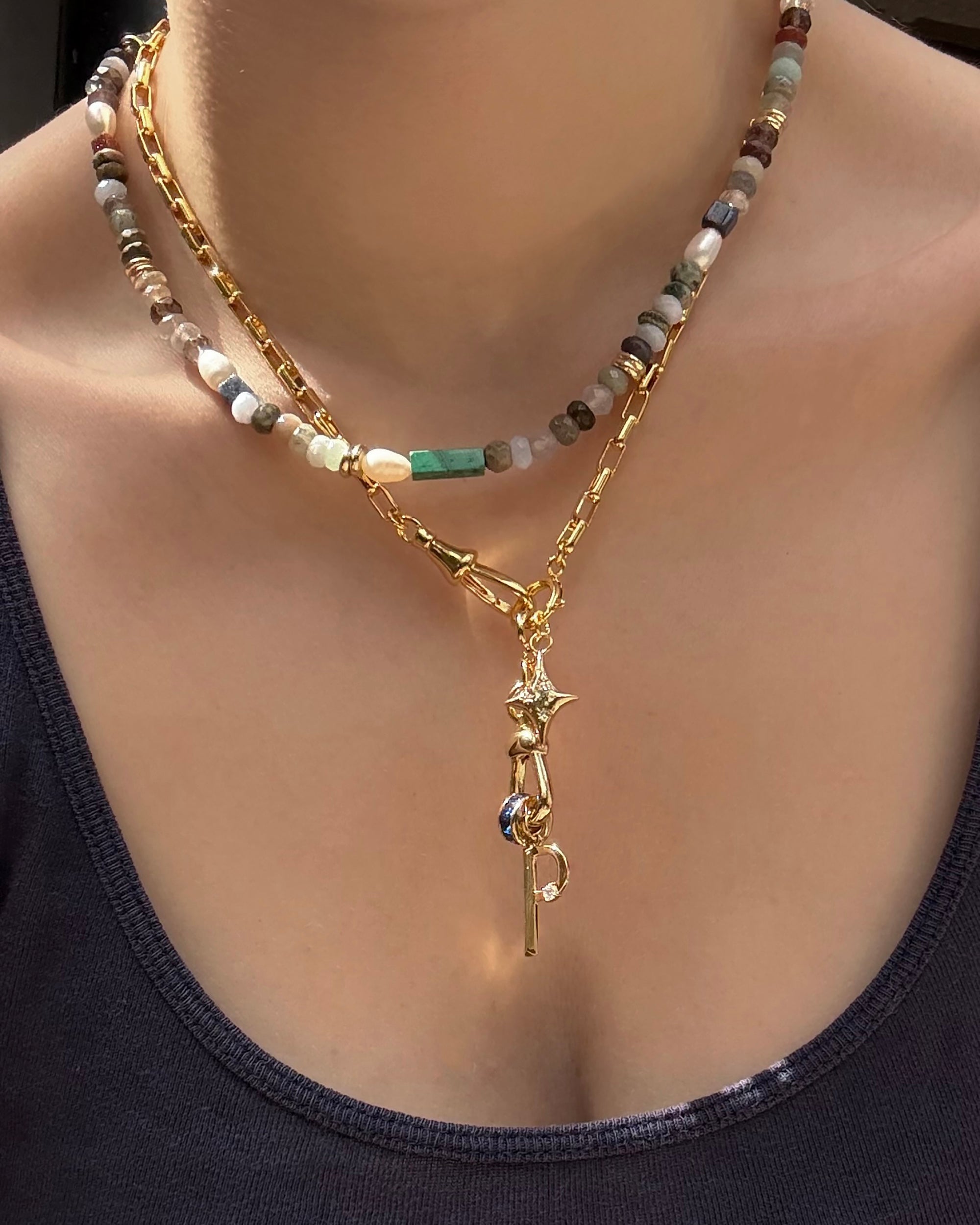 A person wears the Mod + Jo Andy Gemstone Enhancer Chain—multicolored round gemstone beads layered with a gold chain featuring star and key charms—paired with a dark scoop-neck top.