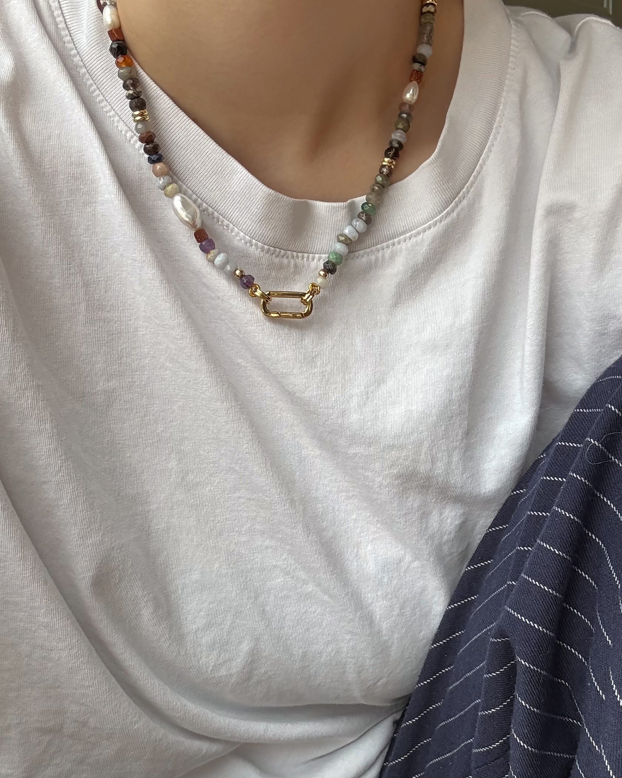 A person wears the Mod + Jo Andy Gemstone Necklace, featuring genuine gemstones and a gold clasp, paired with a white t-shirt. Their arm rests on navy-blue pinstriped fabric in their lap.