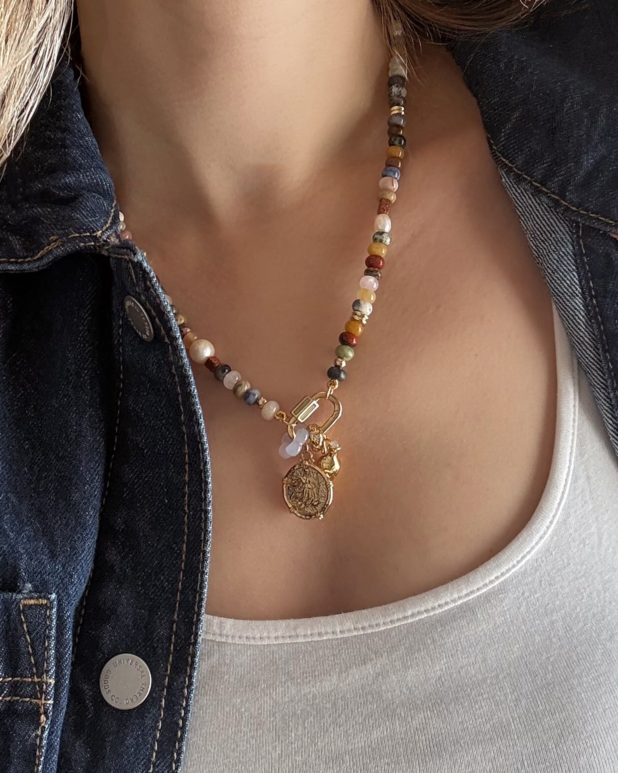 A person wearing the Mod + Jo Juno Flower Gemstone Spacer necklace with authentic gemstones and gold pendants, styled with a white tank top and a dark denim jacket.