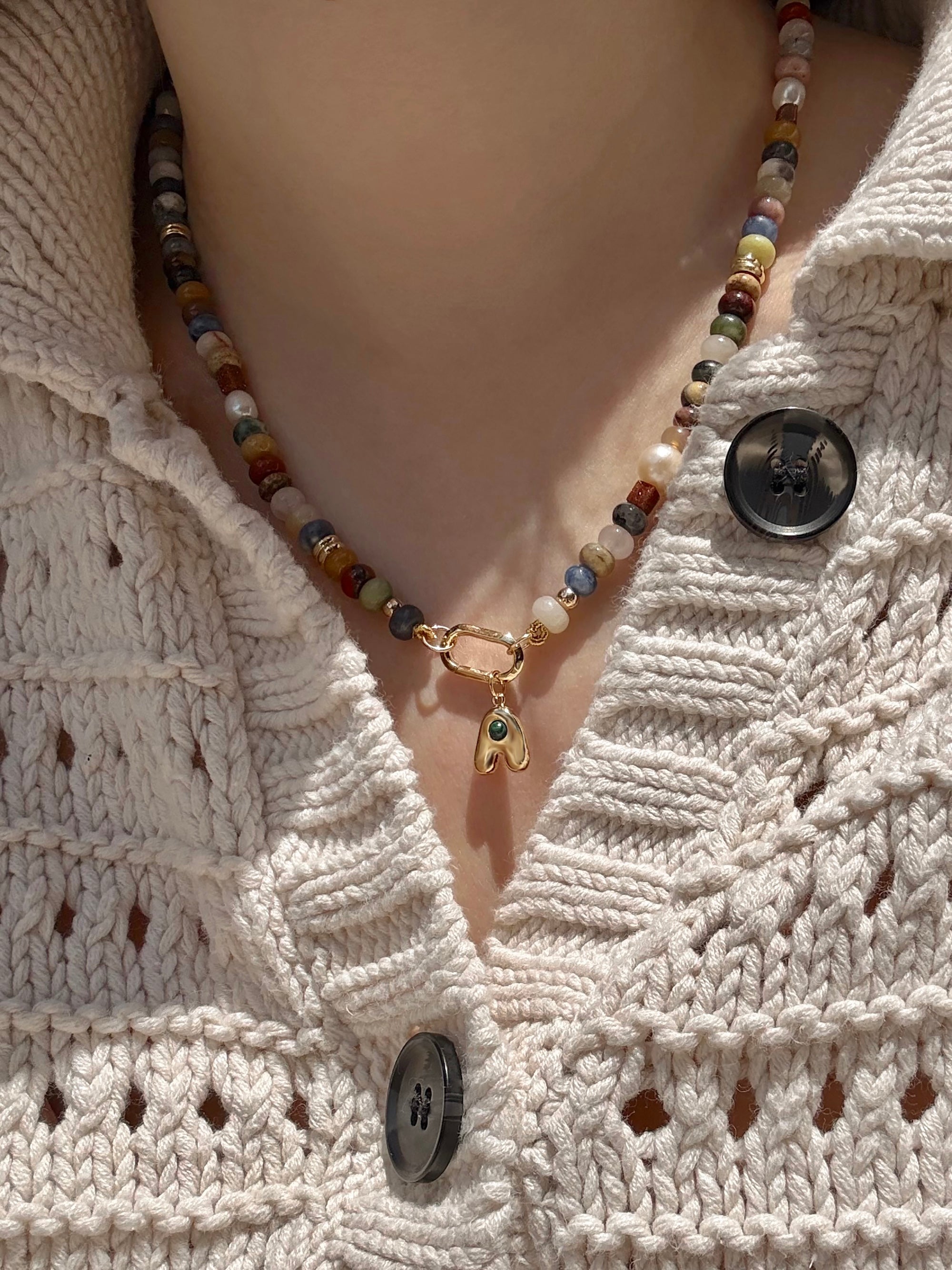 A close-up of a person wearing Mod + Jo’s Thea Bubble Letter Charm necklace over a cream-colored knitted sweater with large black buttons.