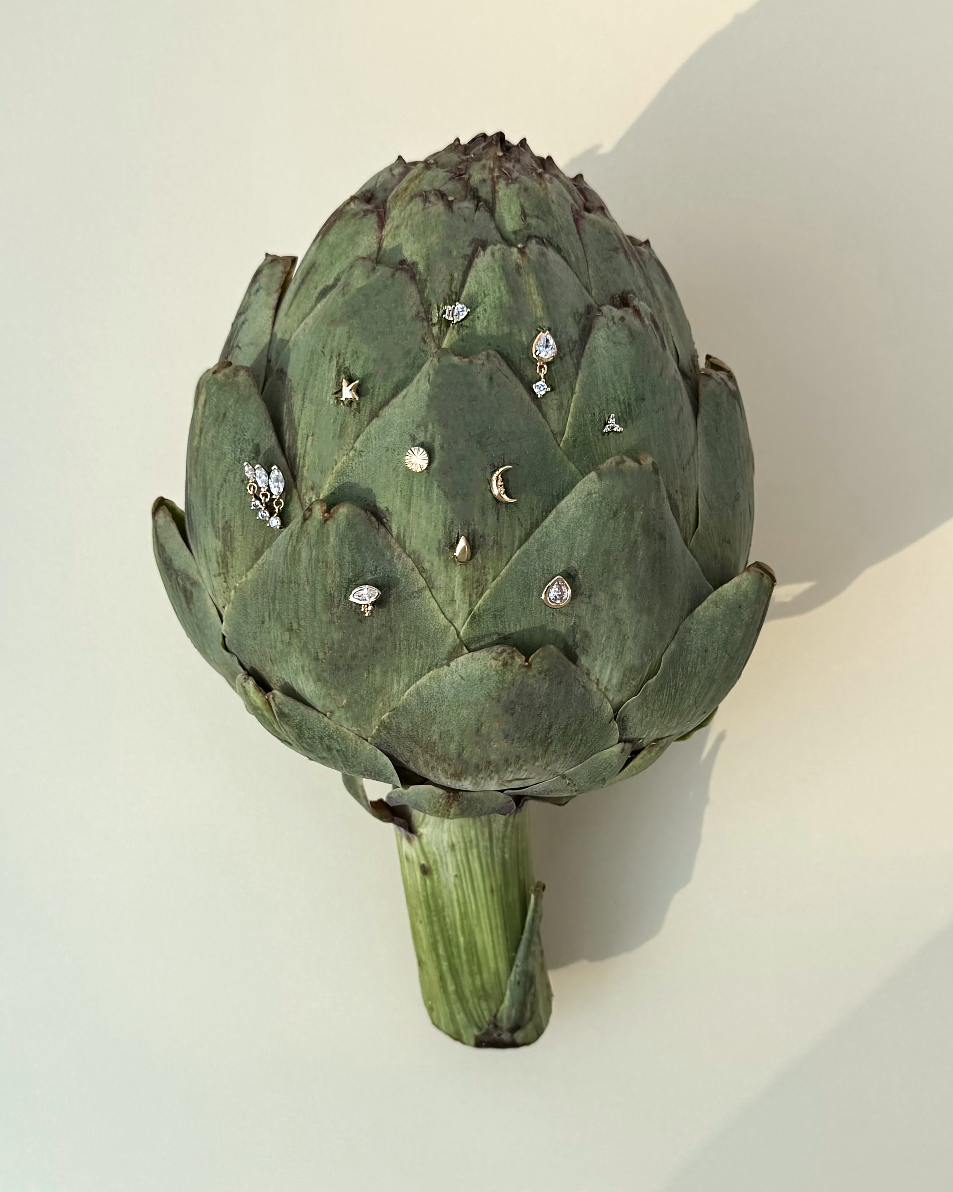 A green artichoke displays an assortment of small, delicate jewelry—gold earrings, rings, and Mod + Jo’s Uma Teardrop Studs—arranged on its leaves against a light beige background.