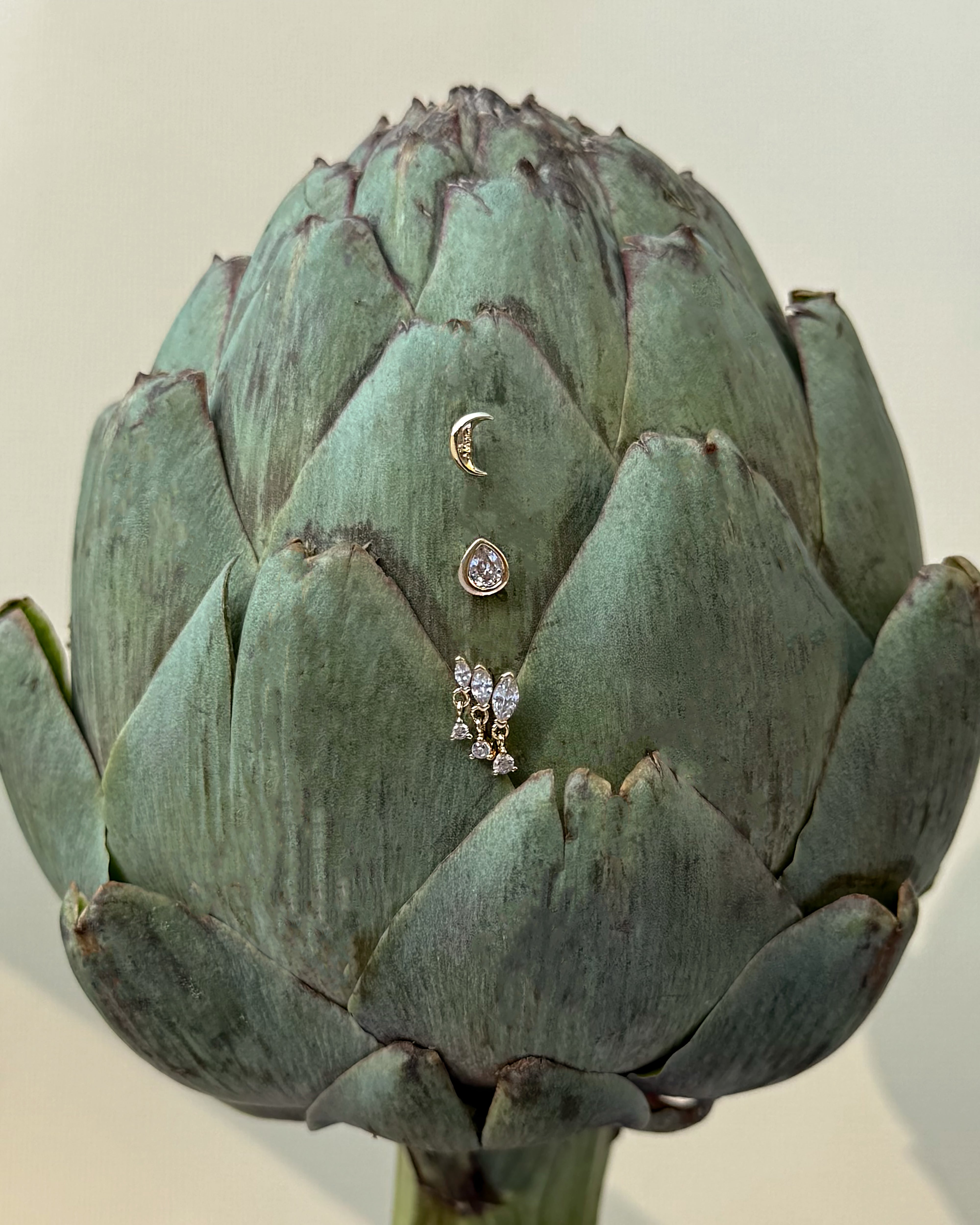 A green artichoke displays three Mod + Jo Gabby Marquise Dangle Studs—each featuring a gold plated crescent moon, a round cubic zirconia gem, and a butterfly—arranged vertically on its front against a plain background.