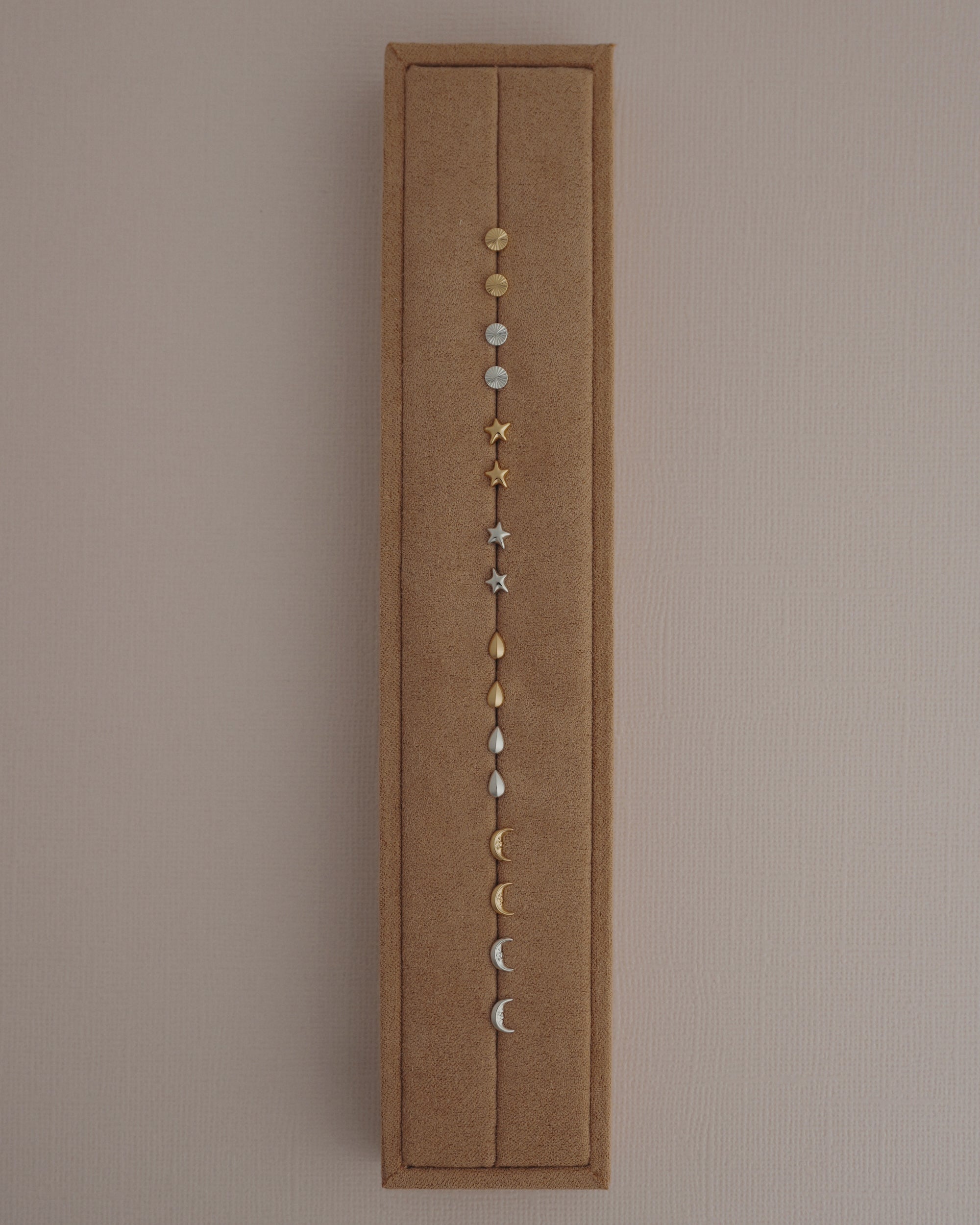 A brown rectangular jewelry display showcases the Mod + Jo Uma Teardrop Studs, along with other small stud earrings in gold and rhodium plated silver, featuring circular, star, teardrop, and crescent moon shapes. These high-shine earrings are arranged vertically against a neutral background.