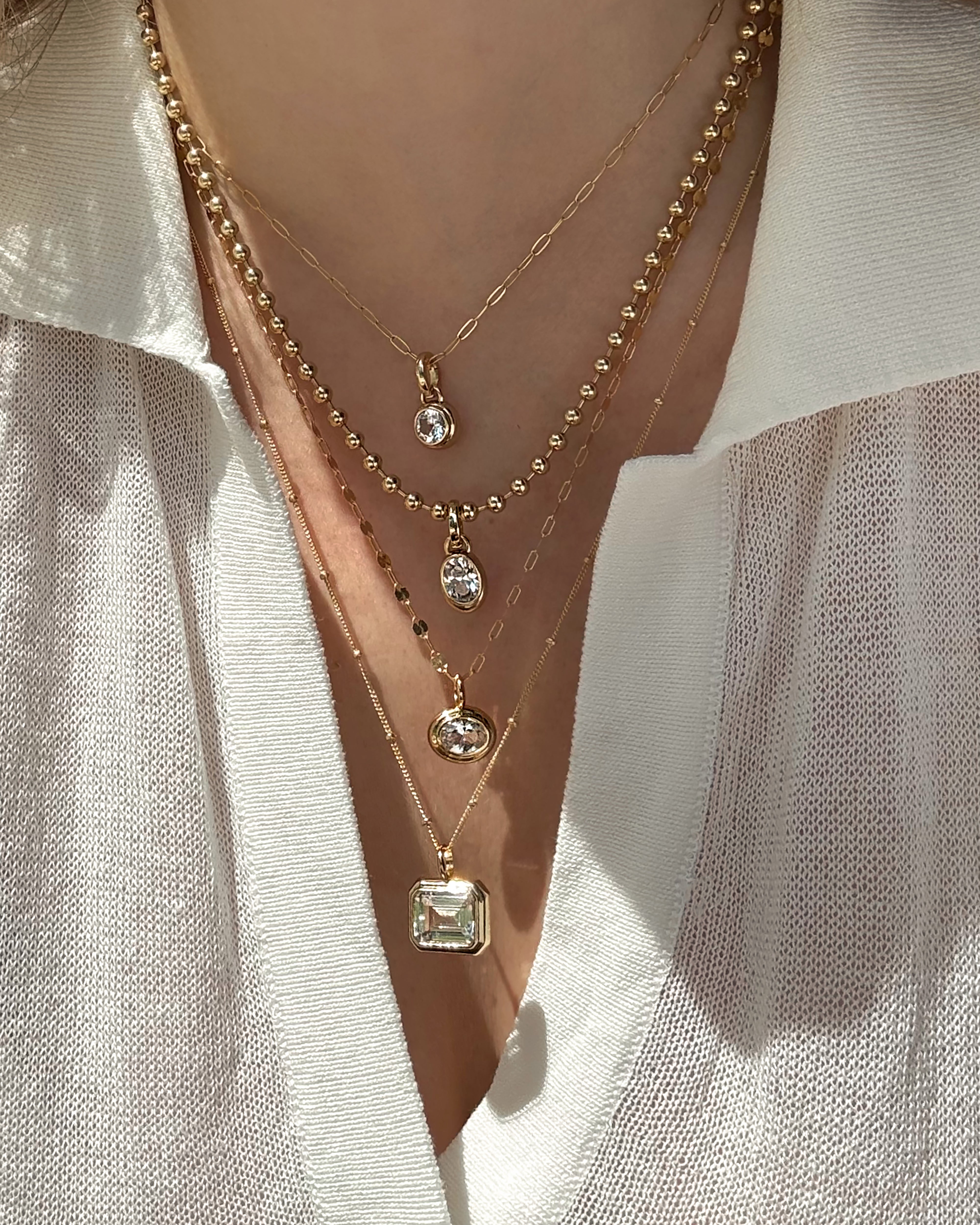 A person wearing a cream v-neck blouse with four layered gold necklaces, including Mod + Jo’s *Preorder* Elsie Sapphire Bezel Charm—an emerald-cut white sapphire pendant sparkling among other gemstones.