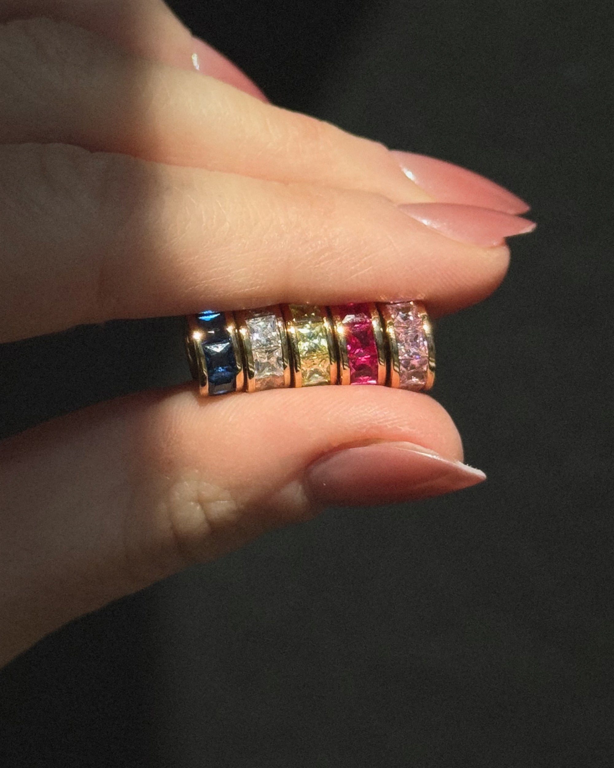A hand with neatly manicured nails holds four gold rings, each with square blue, clear, red, and pink gemstones—perfect to pair with Mod + Jo’s Este Birthstone Charm Spacer—displayed against a dark background.