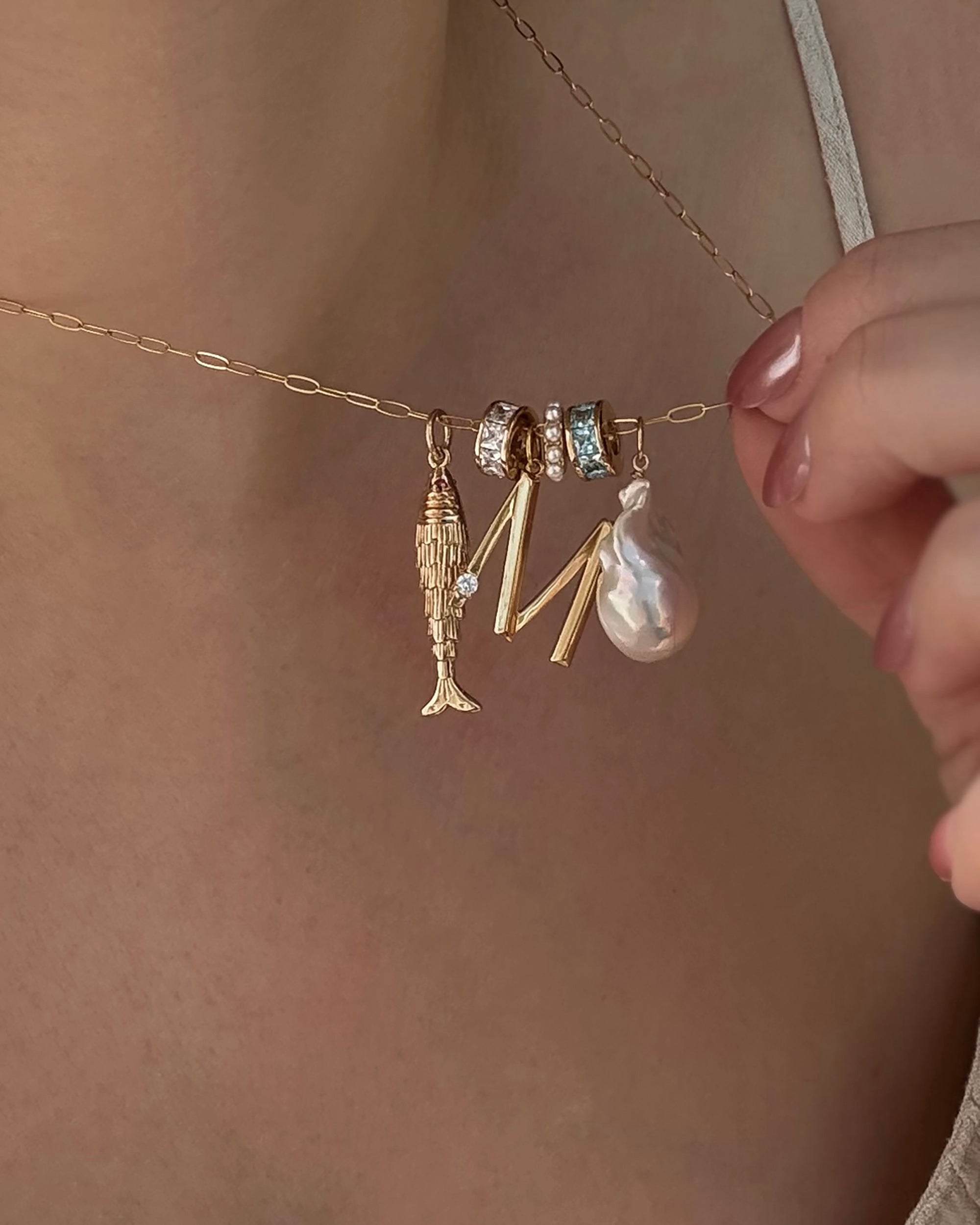 A person with light pink nail polish wearing a thin strap top holds a gold Mod + Jo necklace featuring four charms—fish, two jeweled rings, an M, and a large pearl—showcasing the Este Birthstone Charm Spacer for personalized jewelry.