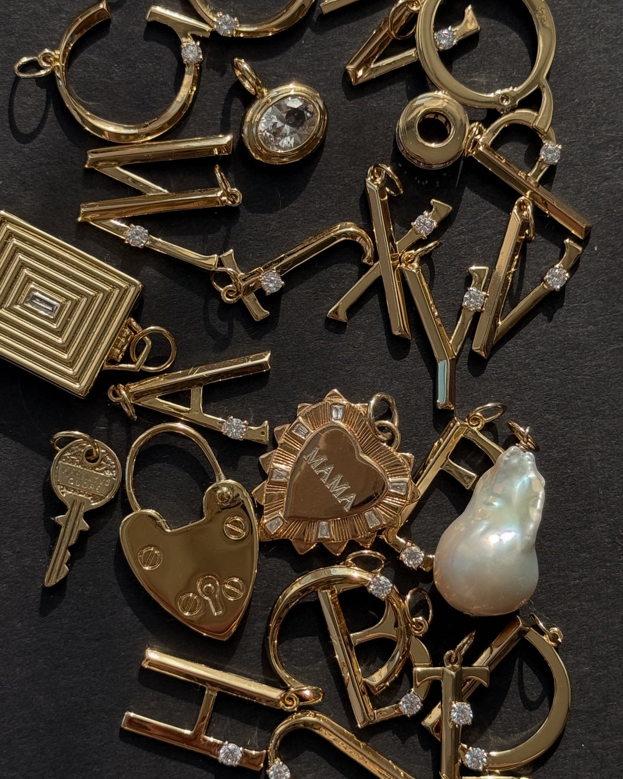 A selection of Mod + Jo charms and pendants, including the Eloise Letter Charm, a heart-shaped lock, key, sunburst “MAMA” pendant, pearl charm, and square locket, displayed on a dark surface.