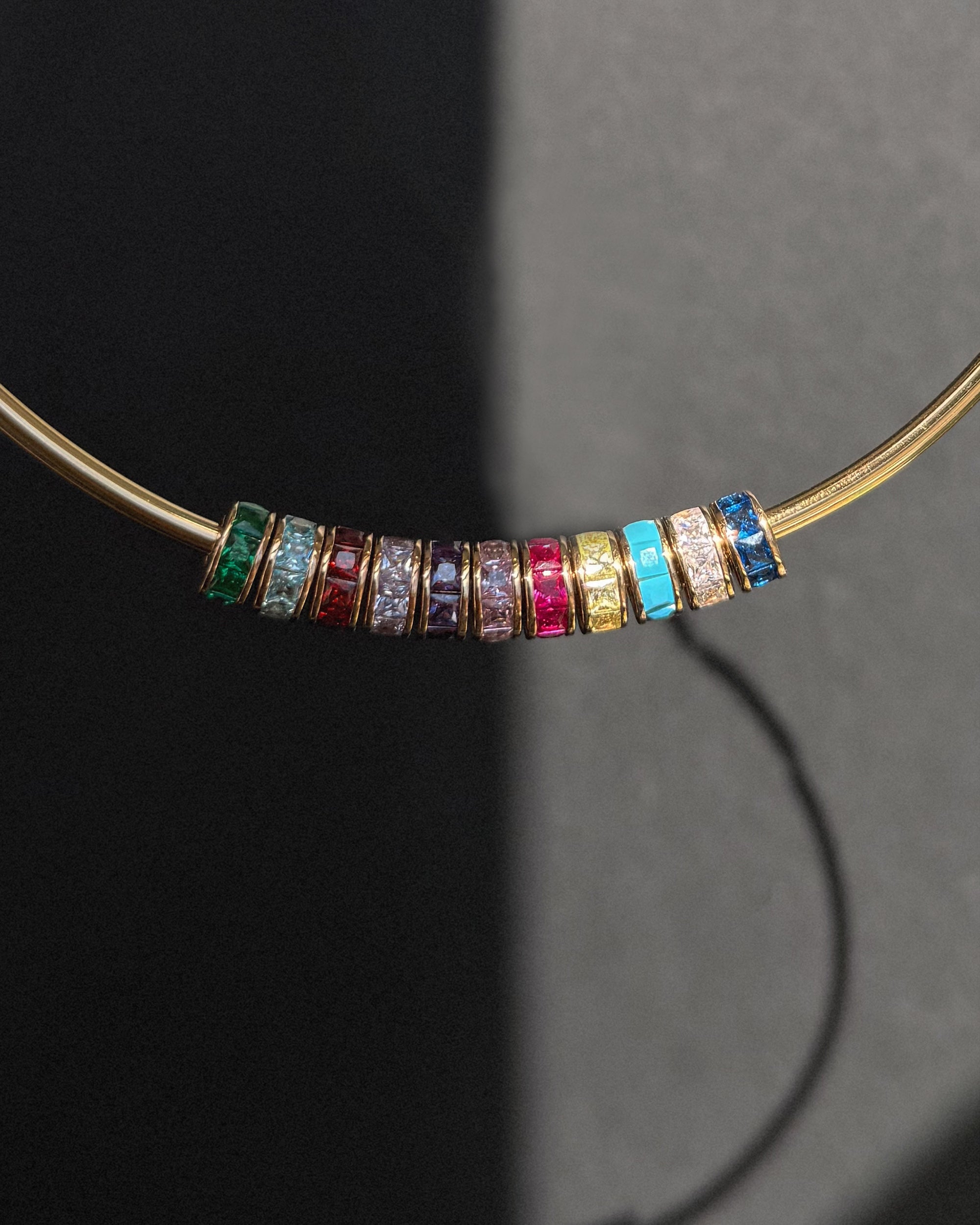 The Mod + Jo Este Birthstone Charm Spacer necklace displays nine colorful gemstone rings, each separated by a slim gold band, elegantly arranged in a row on a dark background with striking shadows.