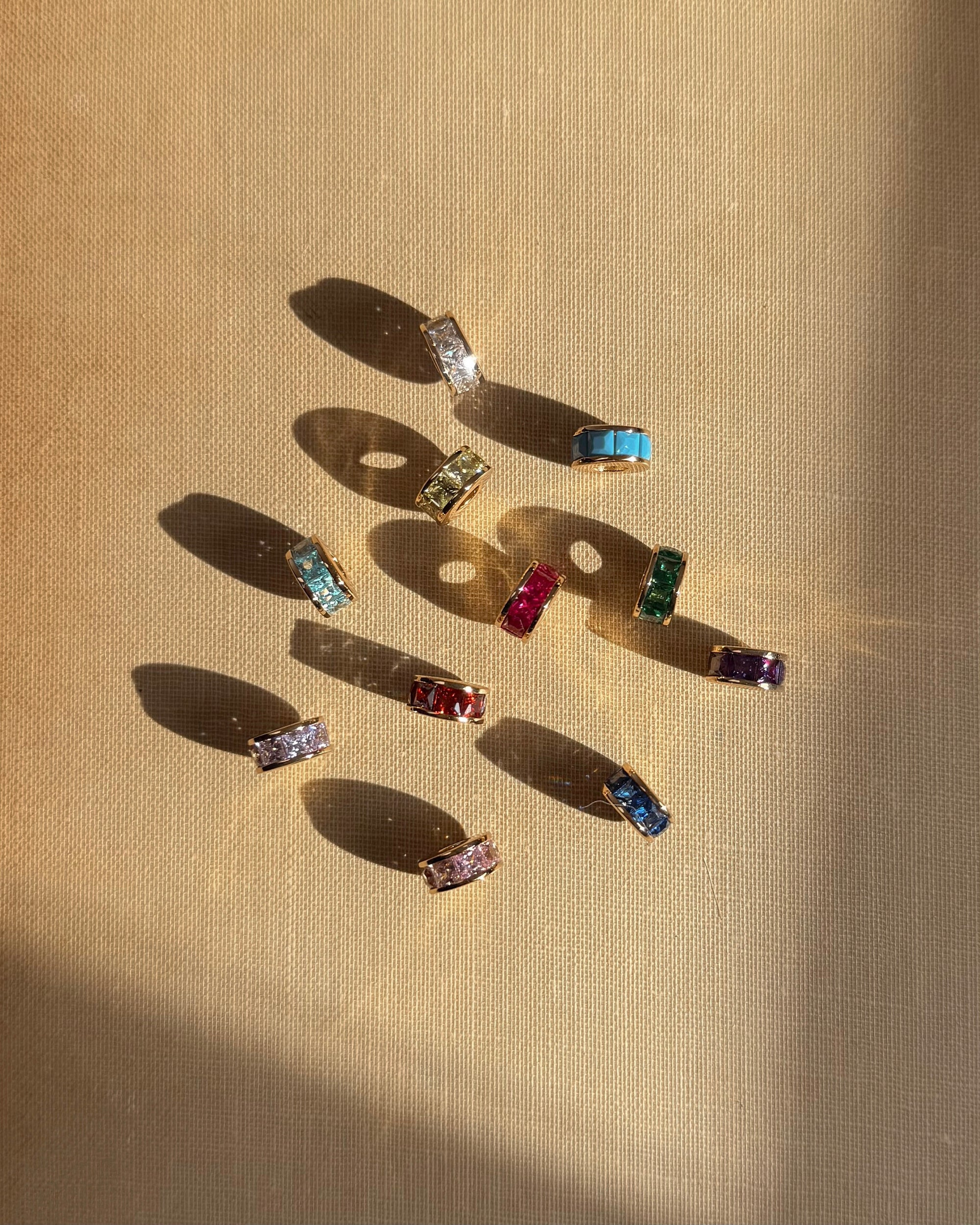 Nine Mod + Jo Este Birthstone Charm Spacer rings, each featuring gold bands and colorful gemstones—blue, green, red, purple, pink, and clear—are displayed on a textured beige surface with sunlight casting unique shadows.